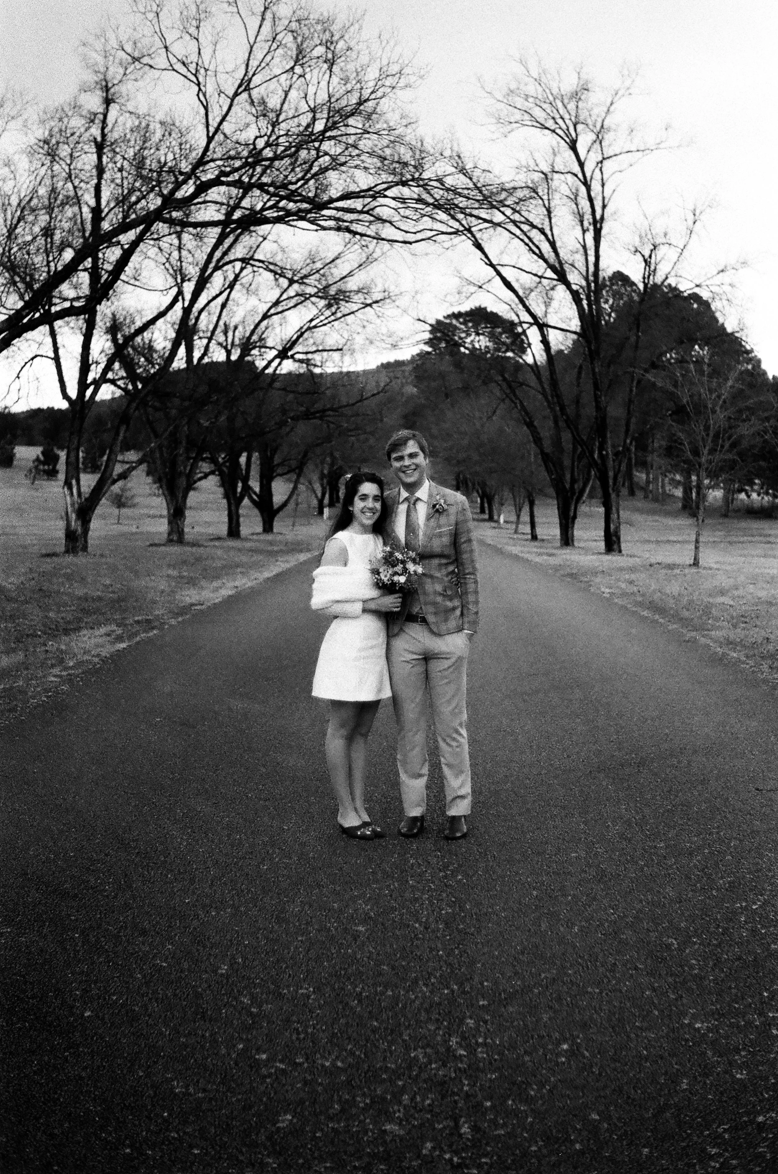 Black and white photograph of a smiling couple standing on a paved road lined with leafless trees, with a distant mountain in the background. The woman is wearing a short dress and holding a bouquet, and the man is dressed in a suit and tie.