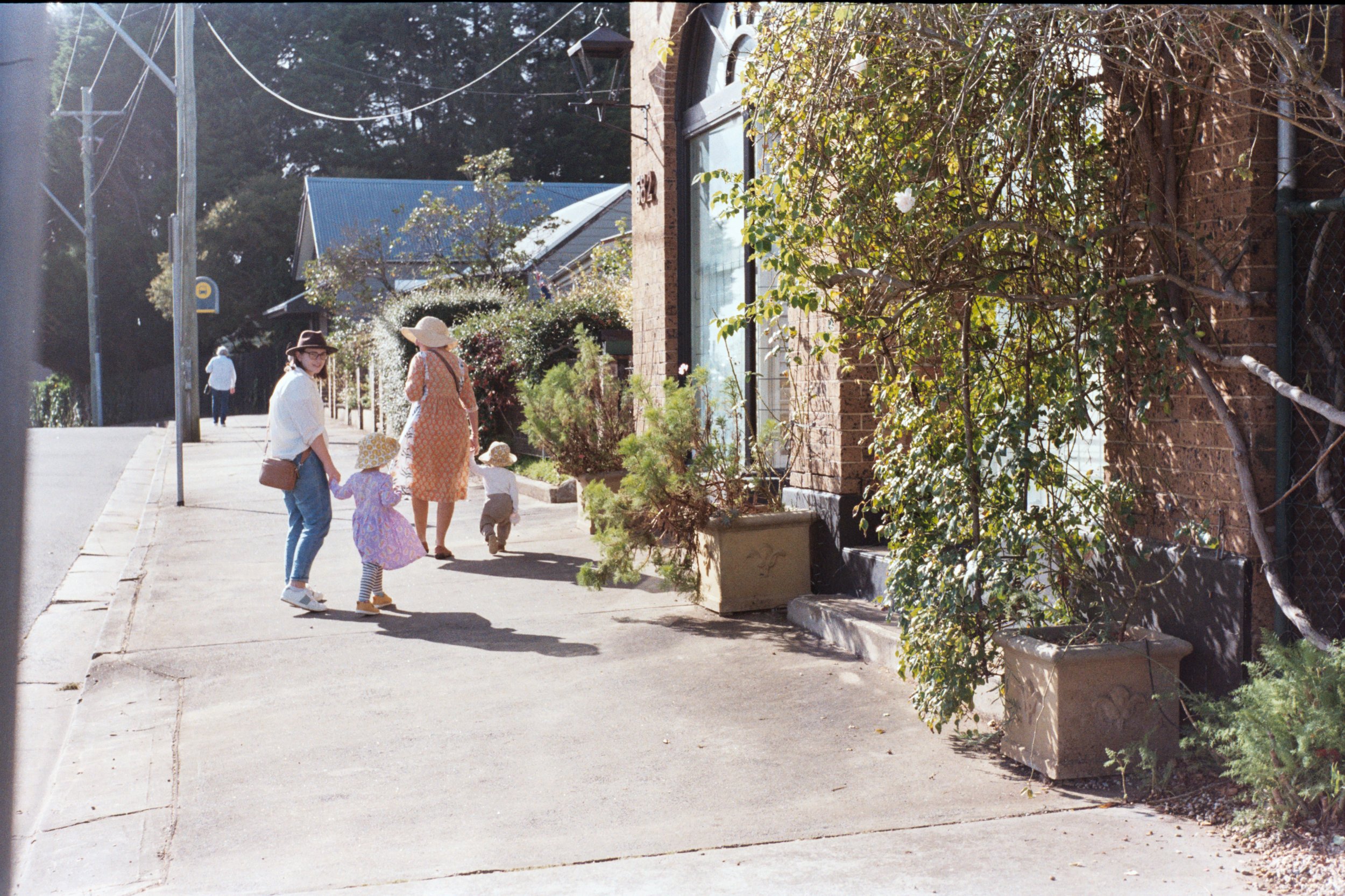 walking on a sidewalk in a suburban neighborhood on a sunny day. The woman is wearing a white top, blue jeans, white sneakers, and a hat, holding hands with a young girl in a purple dress and wide-brimmed hat. The girl is holding the woman's hand. An