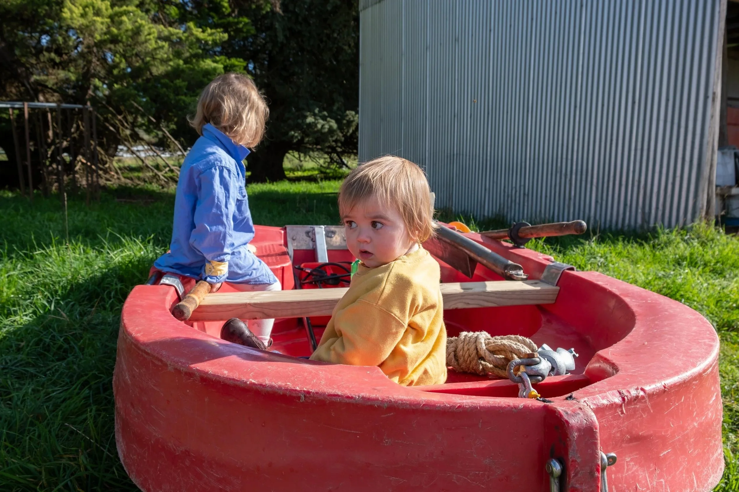 Two young children sitting in a red boat on grass outdoors, with a large tree and a metal building in the background.