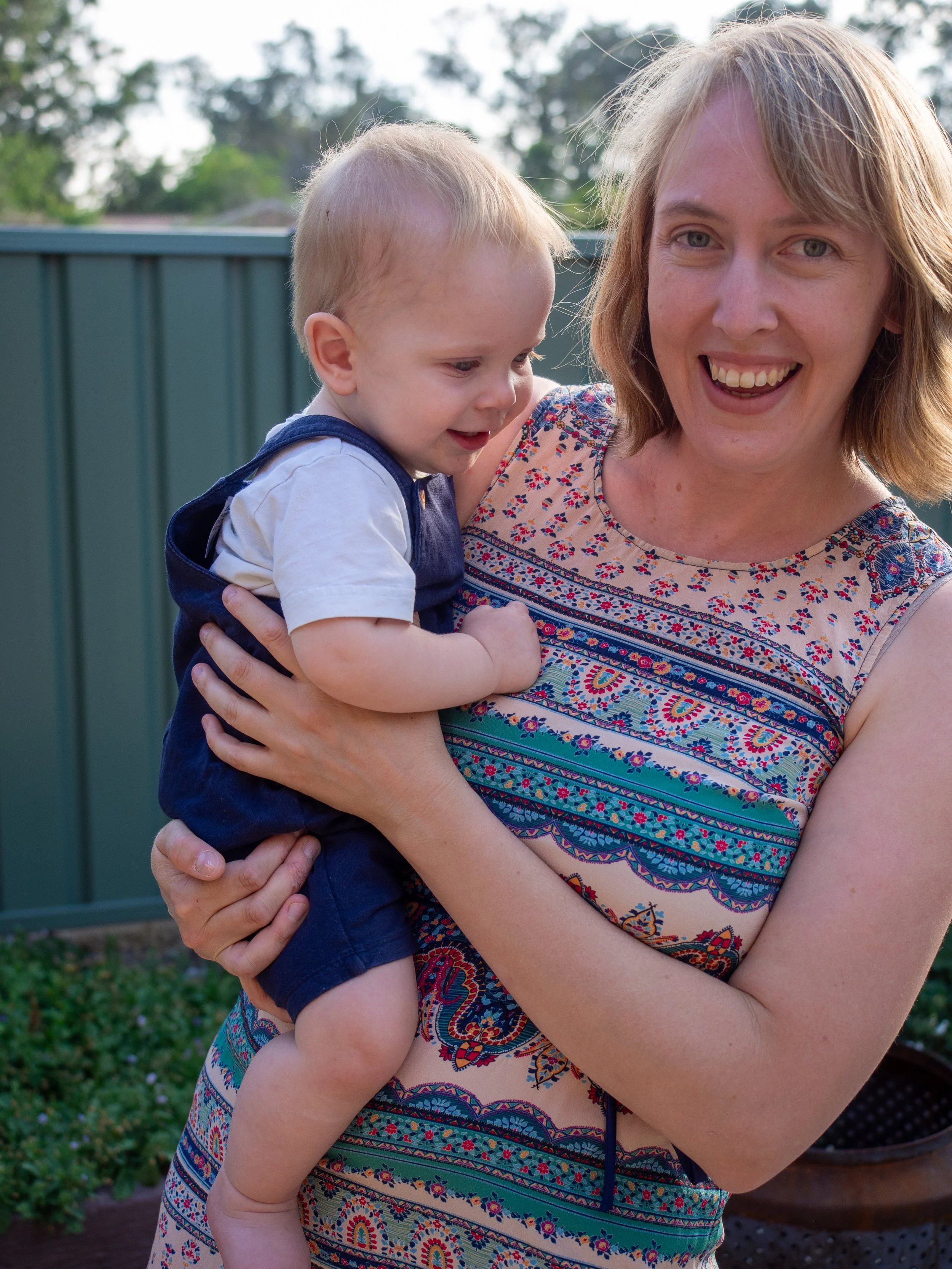 A woman smiling and holding a young child outdoors in front of a green fence and trees.