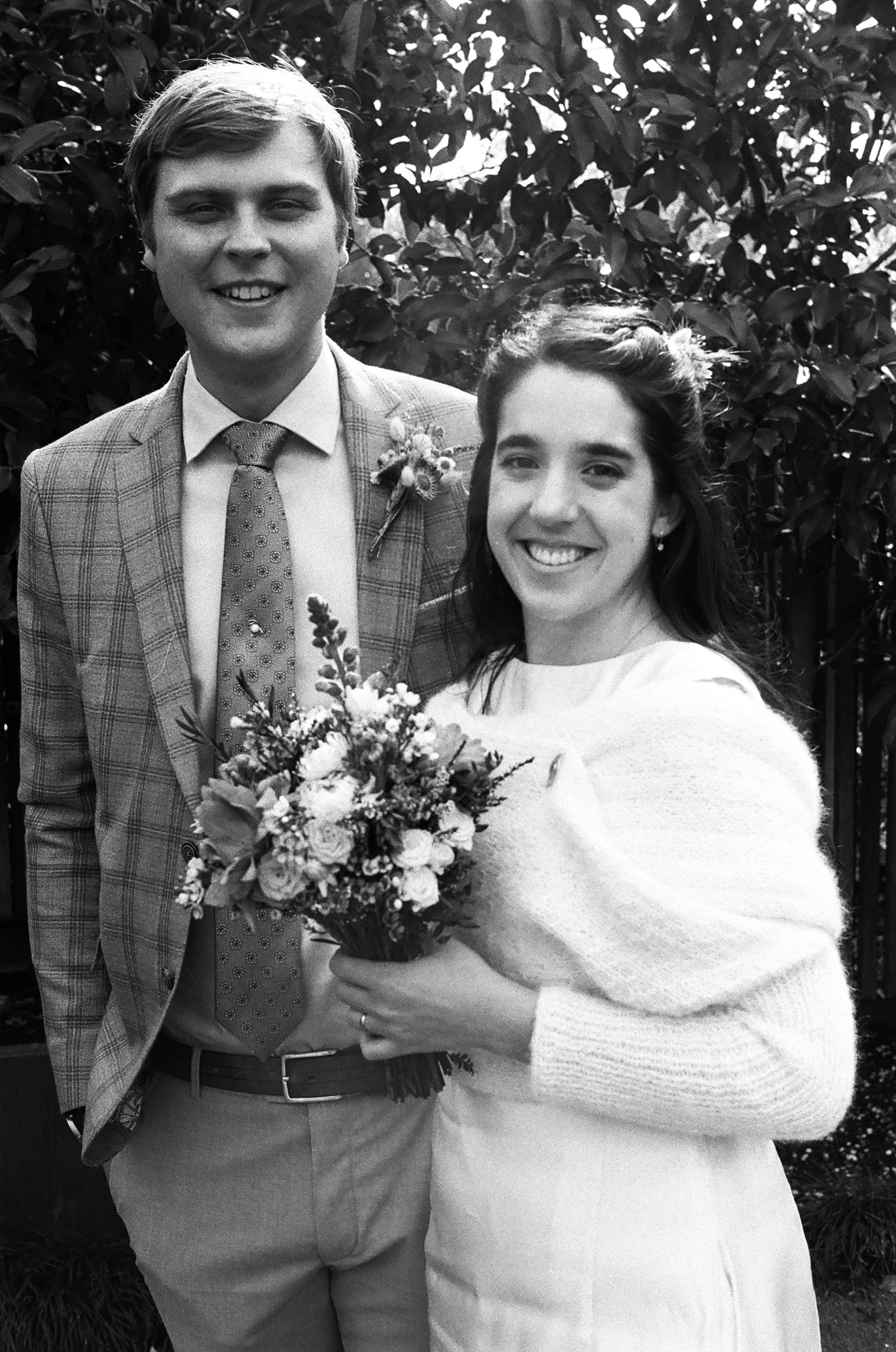 Black and white photo of a smiling couple at a wedding, outdoors with trees in the background. The woman holds a bouquet of flowers and wears a white dress, while the man is in a checkered suit with a boutonniere.