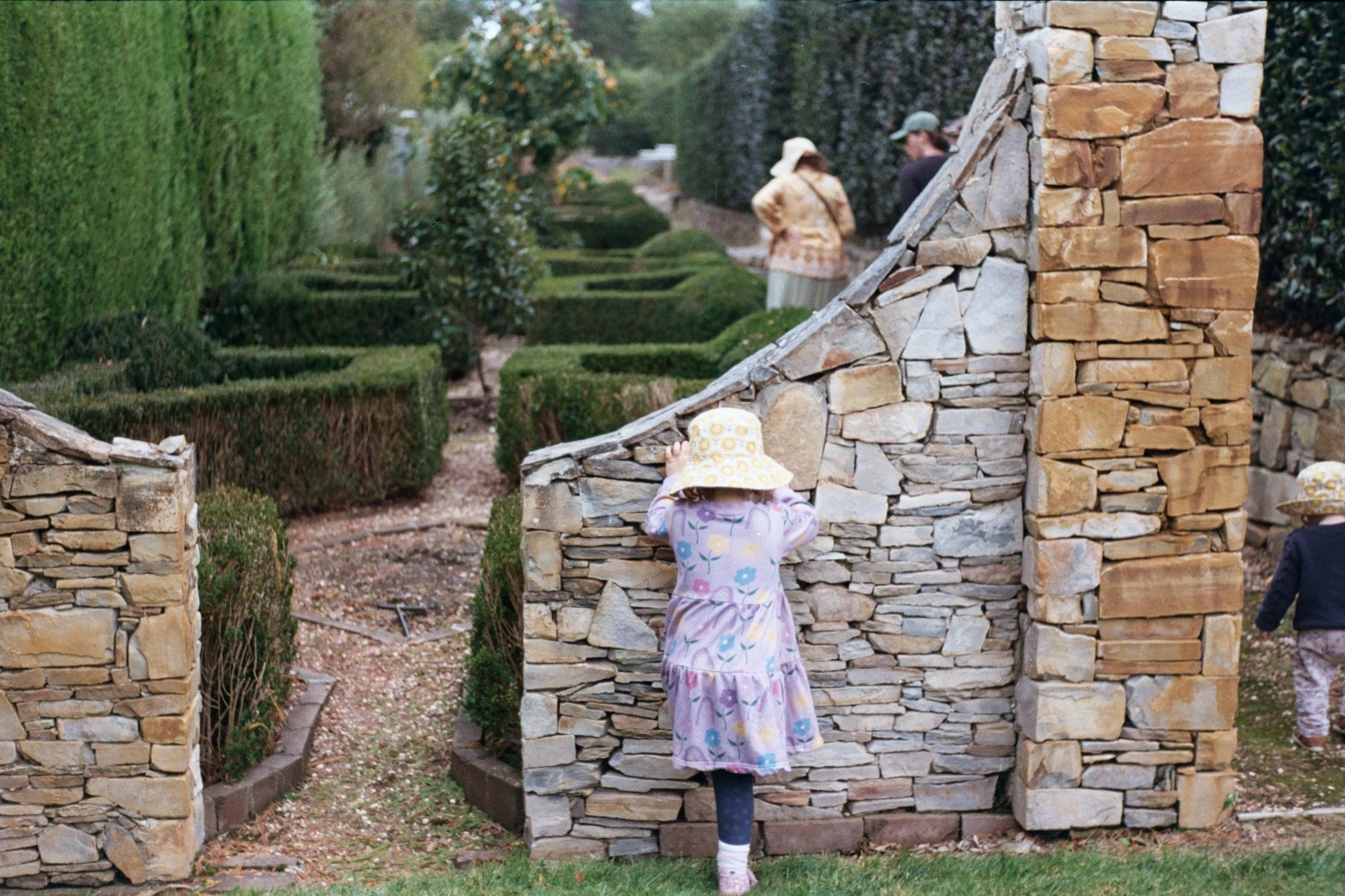 A young girl with a yellow hat and colorful dress looks at a stone wall. Two adults and another child are walking in the background in a garden with neatly trimmed hedges.