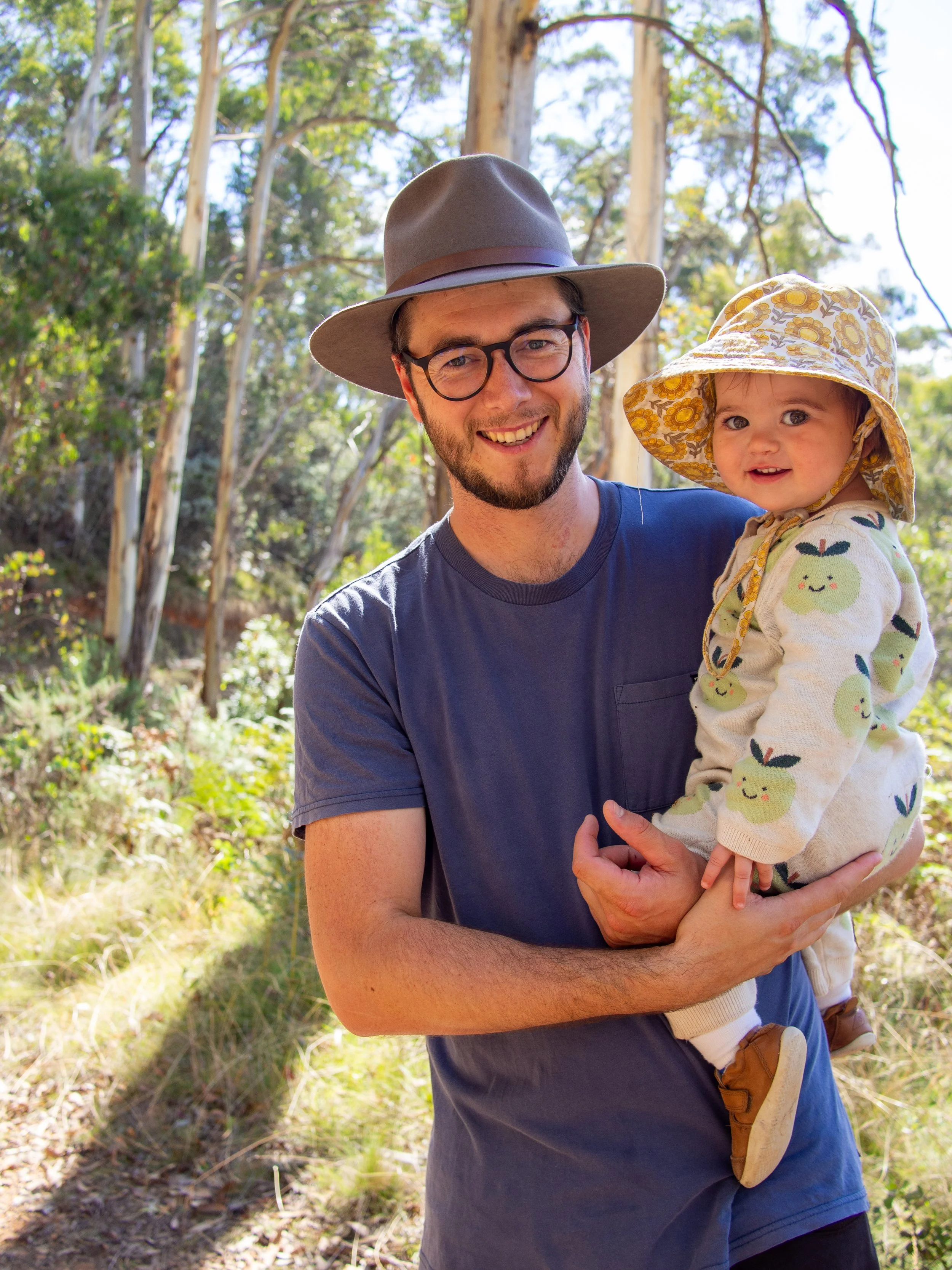 A man wearing a gray hat and glasses is holding a young girl in a forested area with trees in the background. The girl is wearing a wide-brimmed hat and a sweater with a fruit pattern, smiling at the camera.