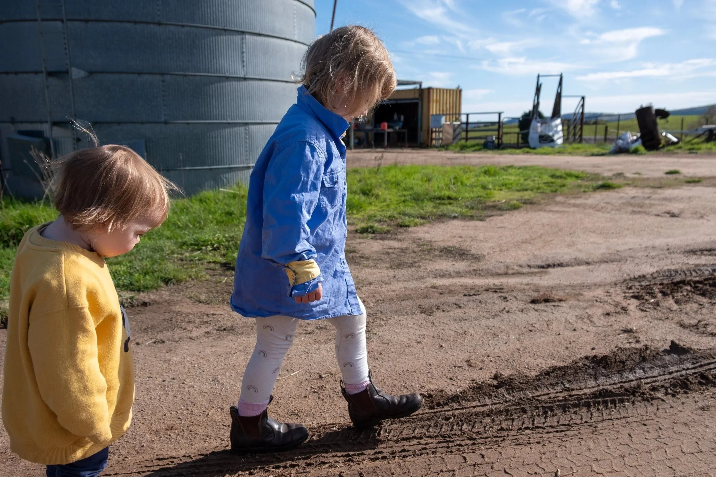 Two young children walking outside on a dirt path near a farm, one in a yellow sweater and the other in a blue jacket, with farm buildings and containers in the background.