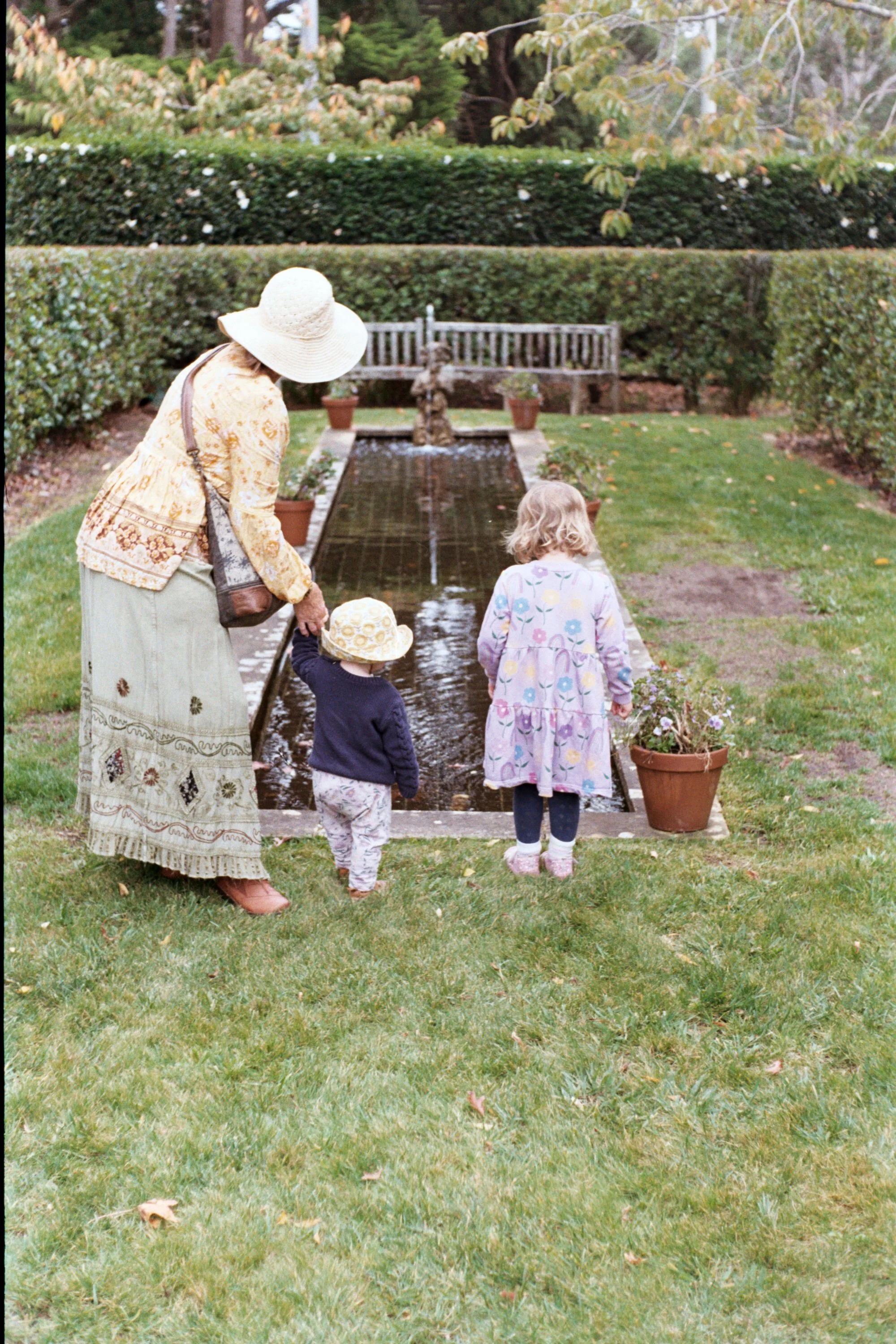 An elderly woman with a wide-brimmed hat and patterned skirt stands with two young children by a narrow garden pond, observing the water.