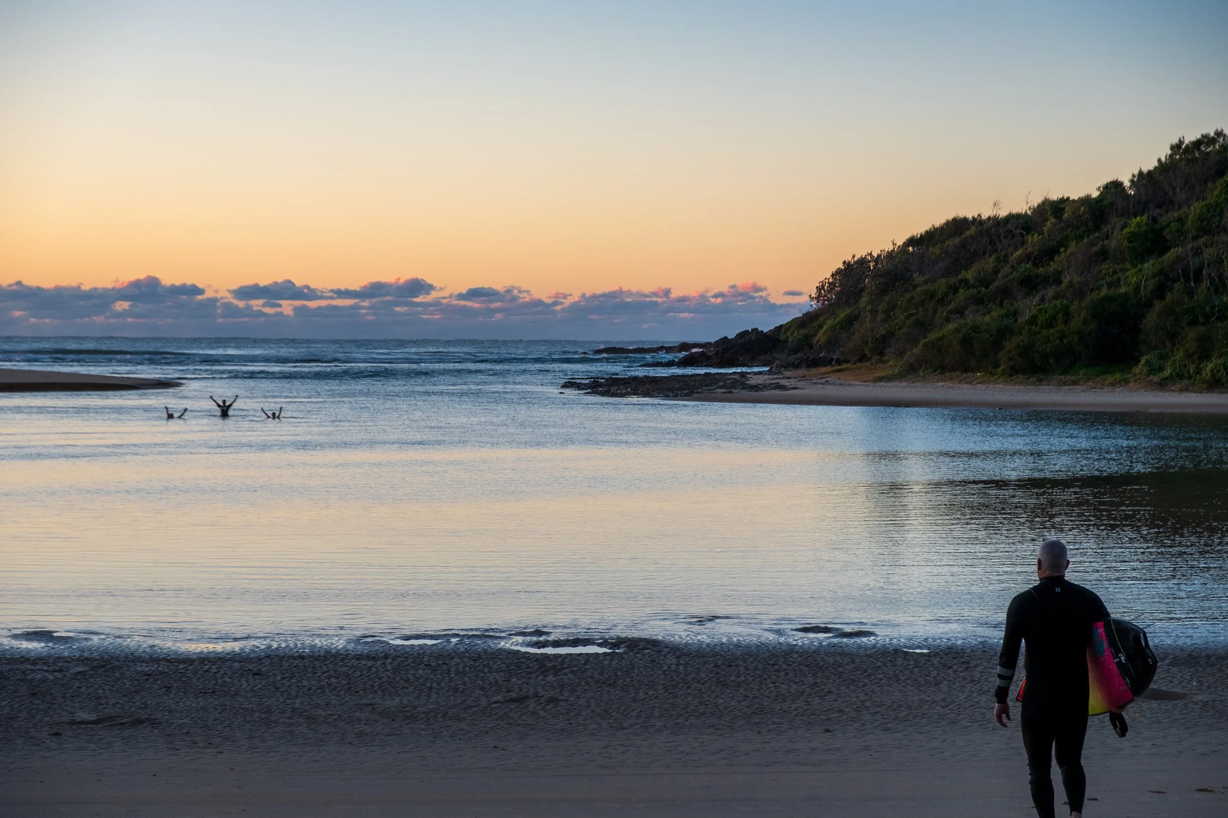 A person carrying a surfboard walking on a beach during sunset with a boat in the distance and a green hillside on the right.