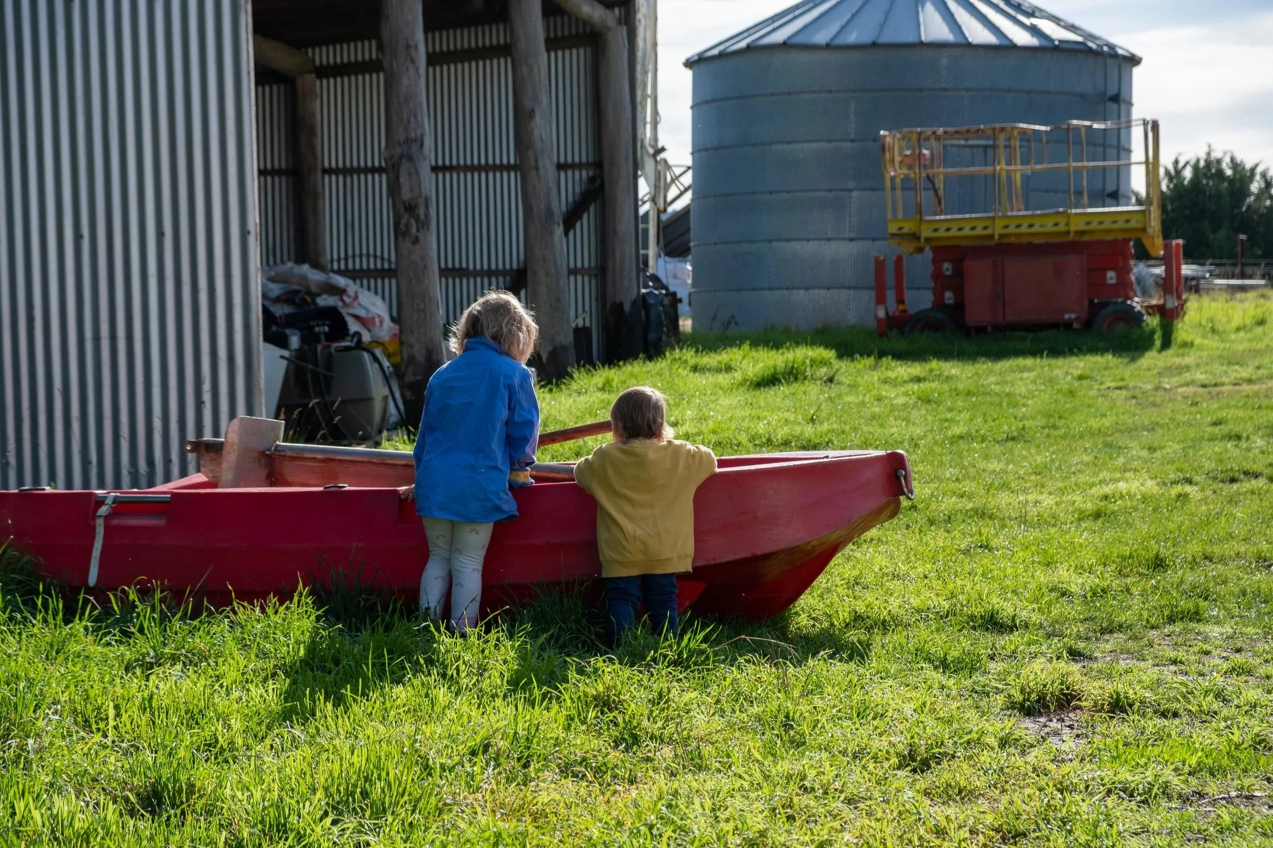 Two children, a girl and a boy, are playing near a red boat on a grassy field in a farm setting. The girl, in a blue jacket and light-colored tights, is standing on the ground, and the boy, in a yellow shirt, is leaning on the boat's edge, looking inside.