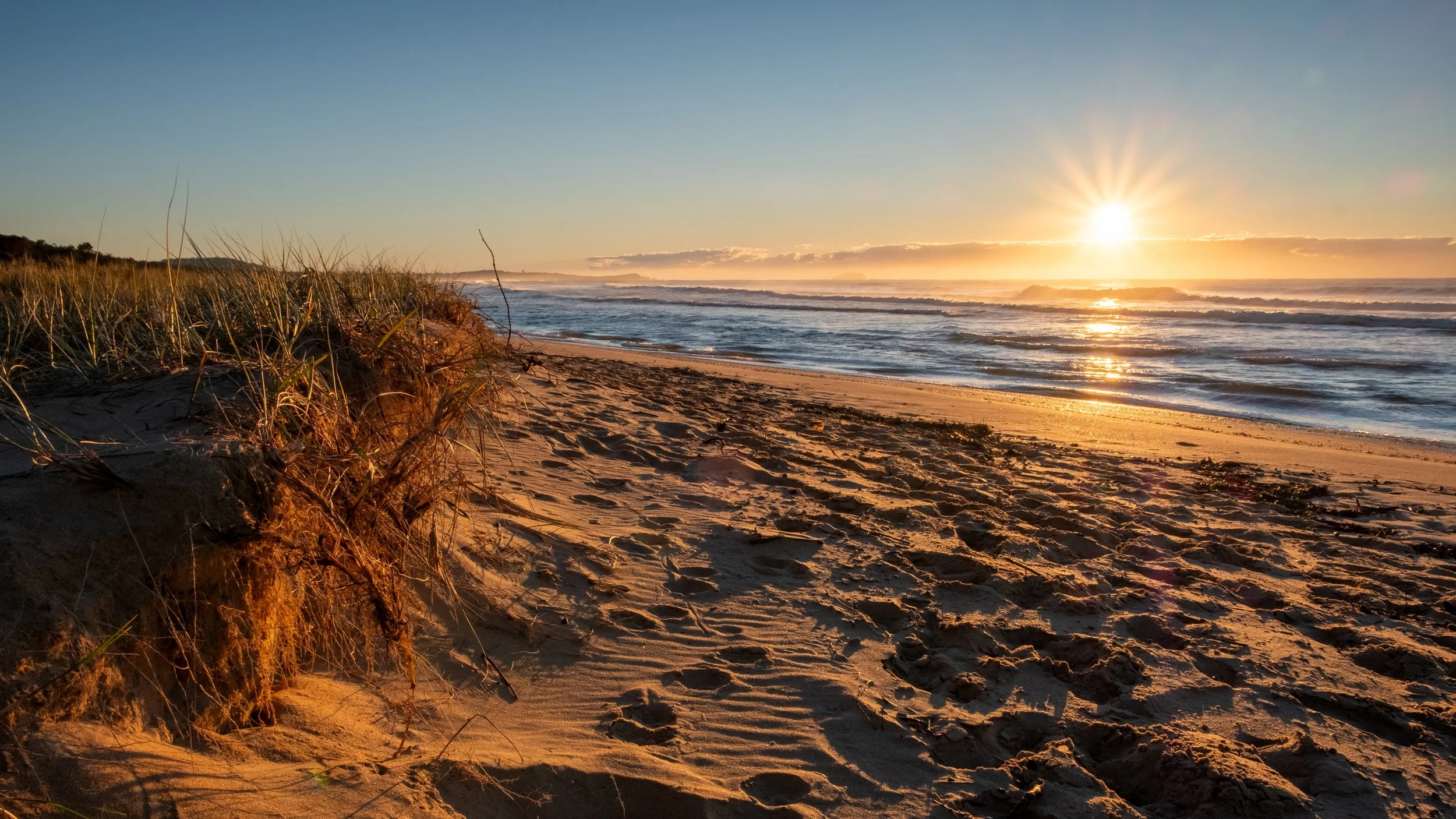 A sandy beach at sunset with grass-covered dunes on the left, gentle waves on the right, and the sun low on the horizon, casting a warm glow over the scene.