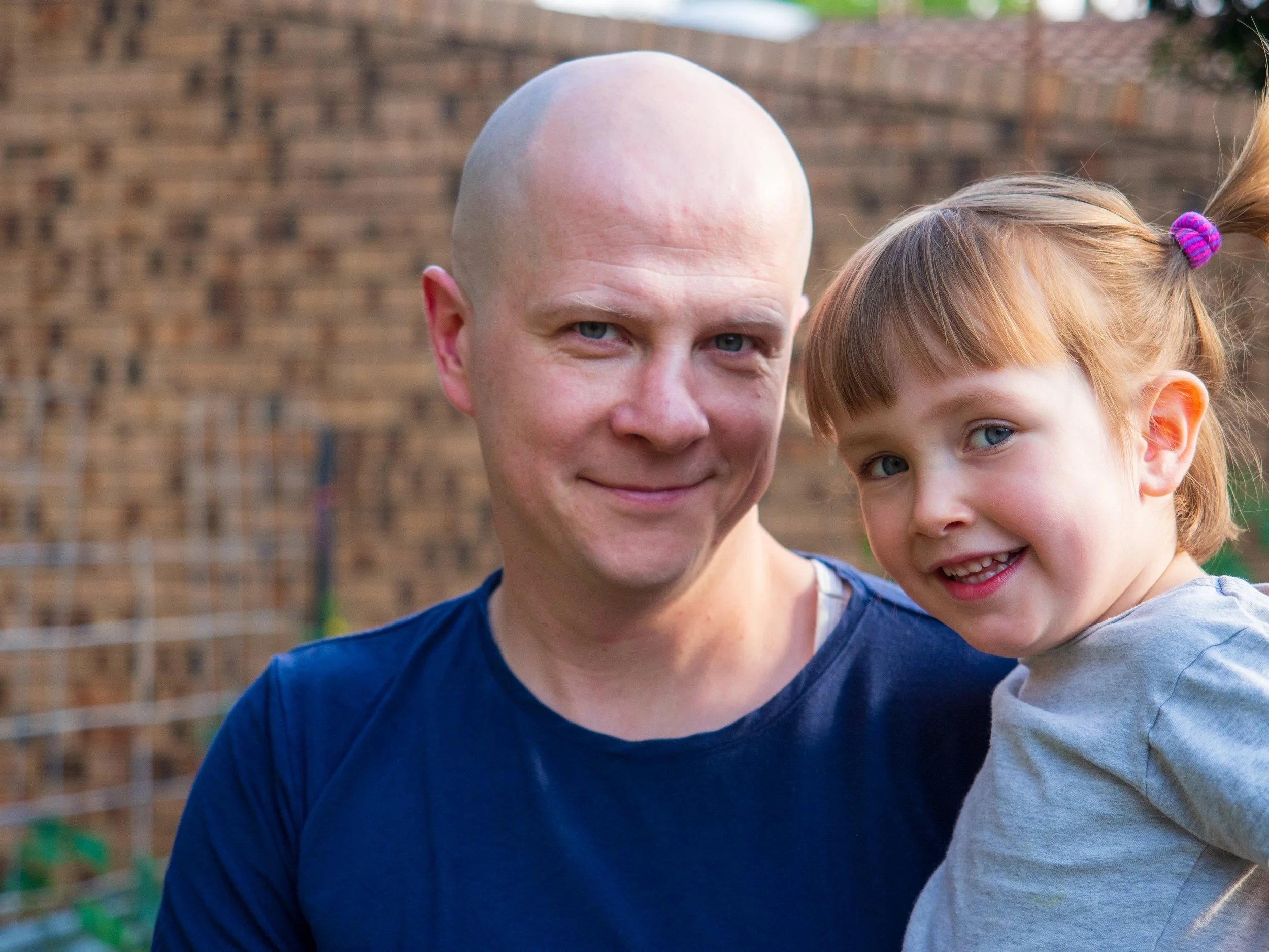 A man with a shaved head and a young girl with red hair tied in pigtails standing outside near a brick wall, smiling at the camera.