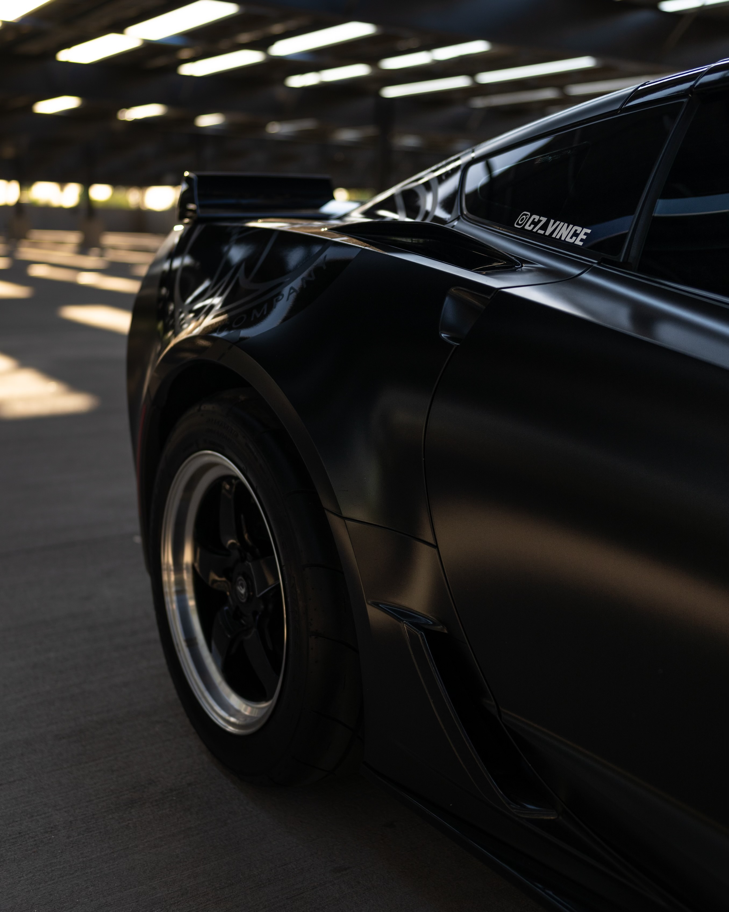 Close-up of a black sports car in a parking garage, showing the front left side with a shiny finish and a tire.