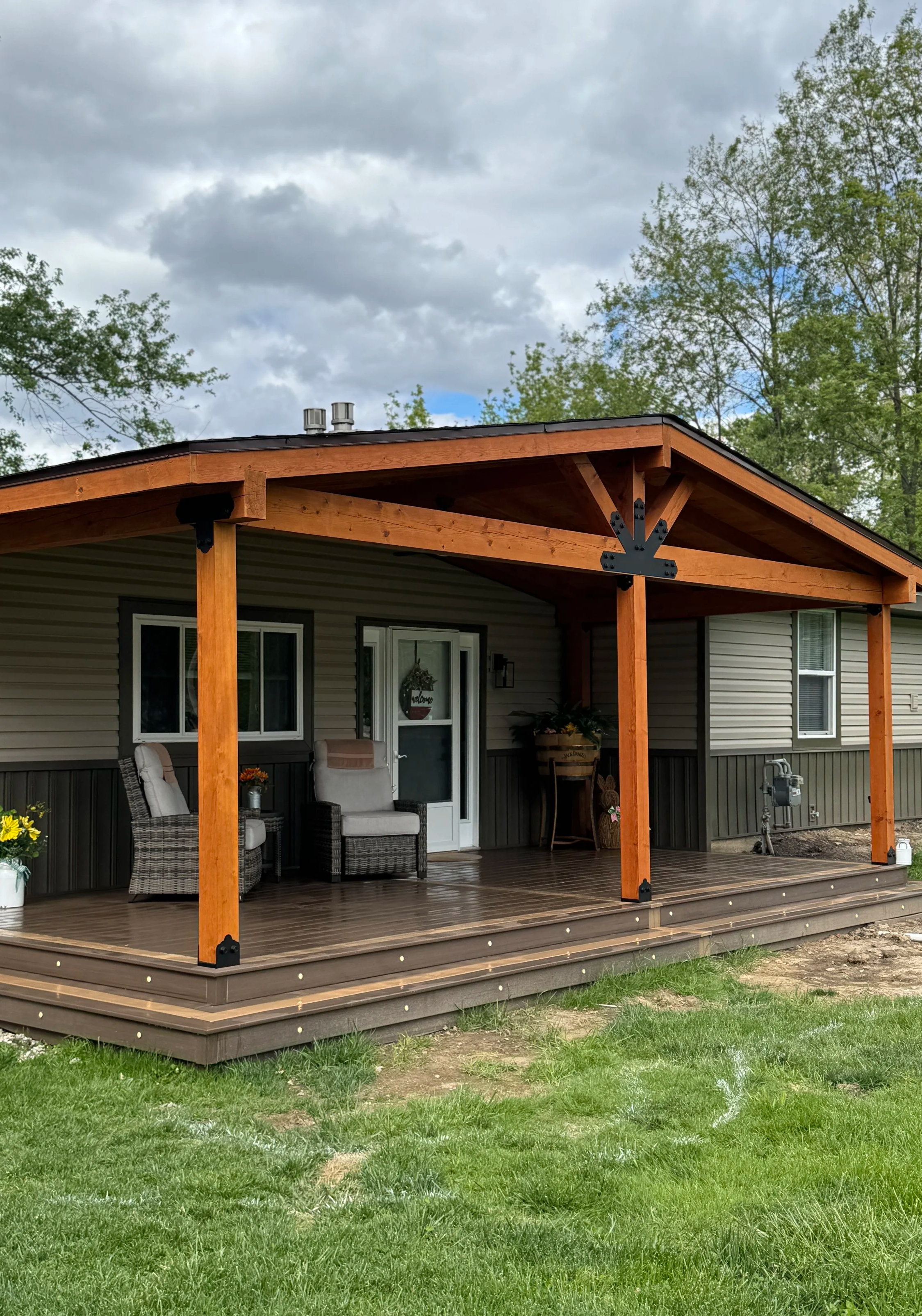 A backyard patio with a wooden covered deck, potted plants, outdoor furniture, a grill covered with a black cover, and string lights hanging from the wooden beams. There are trees in the background.