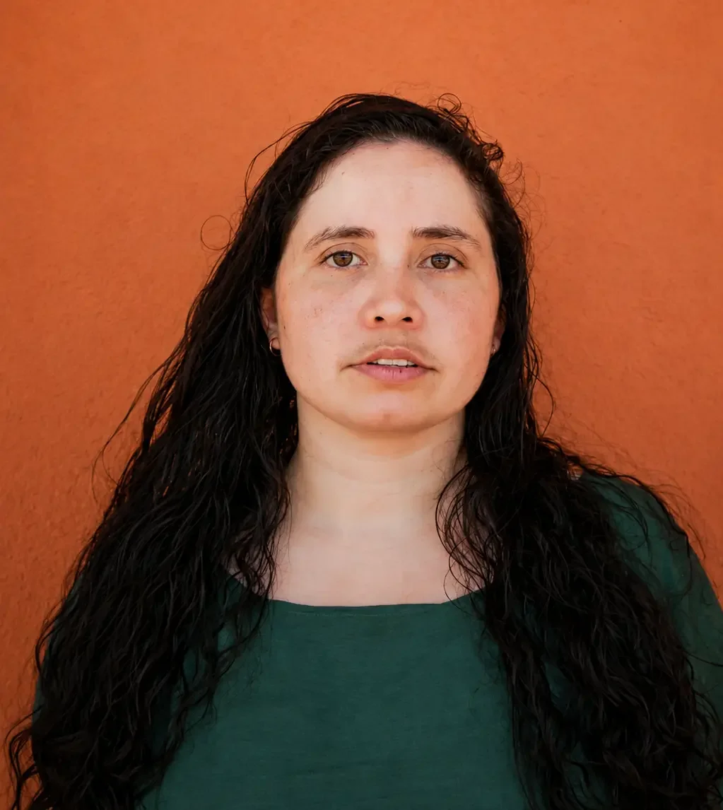 A woman with long black curly hair, wearing a green top, standing against an orange wall.