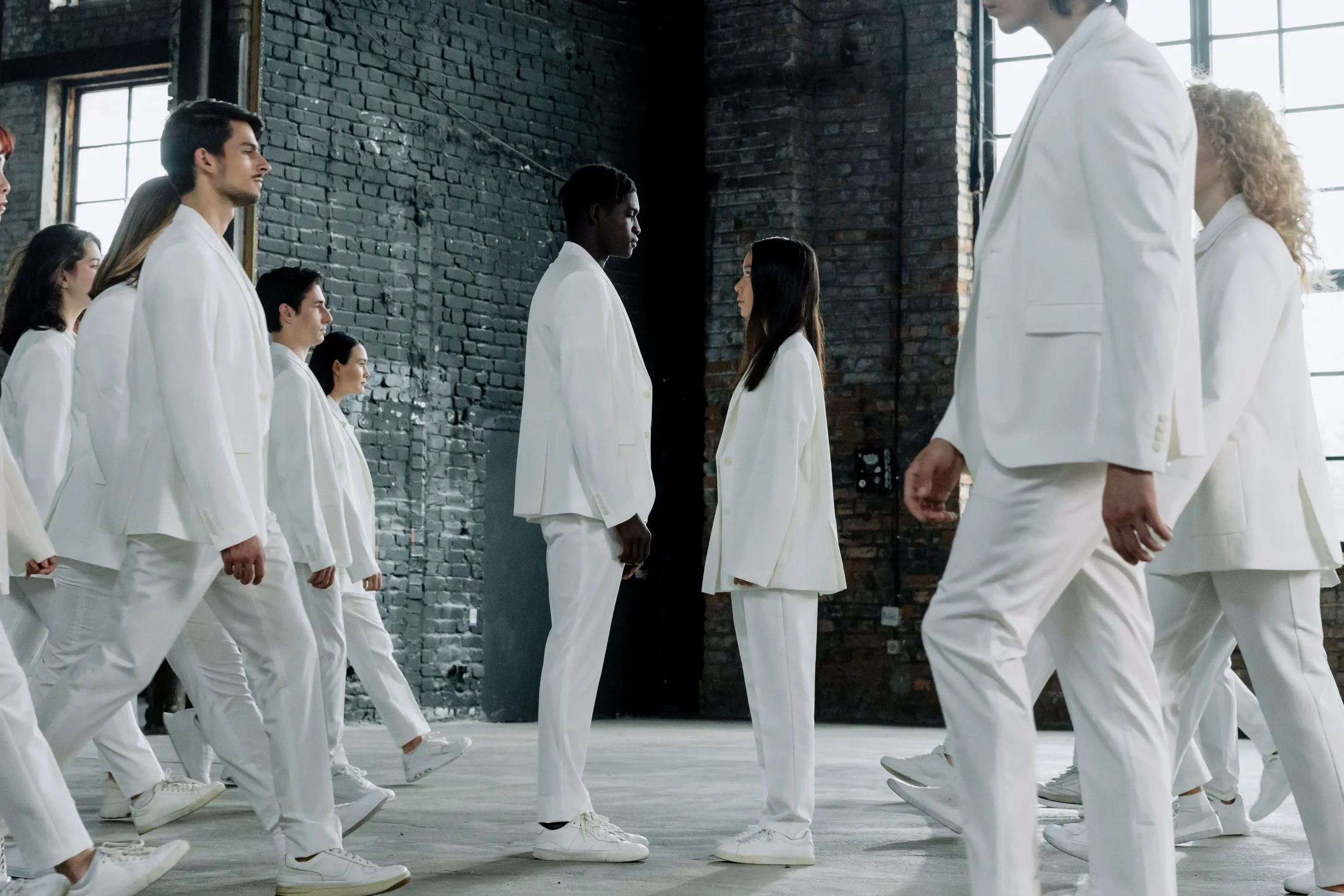 A group of people dressed in white suits standing in a line facing each other in an industrial-style room with exposed brick walls and large windows.