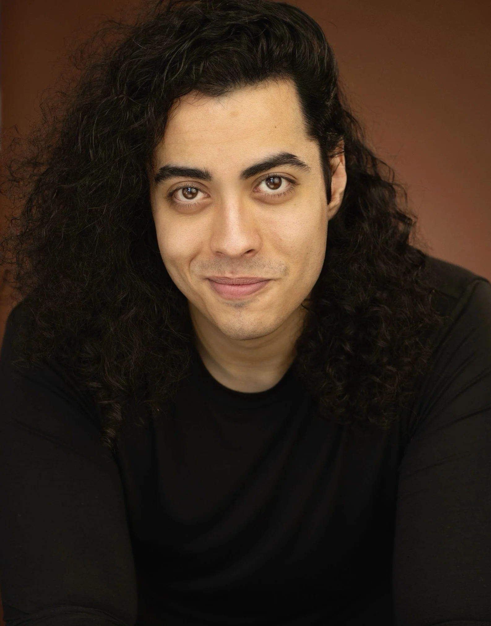 Close-up portrait of a young man with long, curly black hair, wearing a black shirt, smiling subtly, against a brown background.