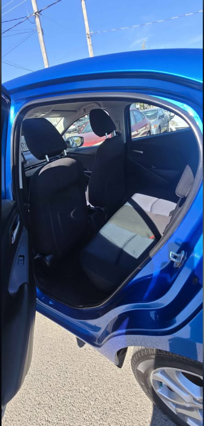 Inside view of a blue compact car showing the back seat area with black seats and headrests, with cars visible through the open door and the blue exterior of the car.