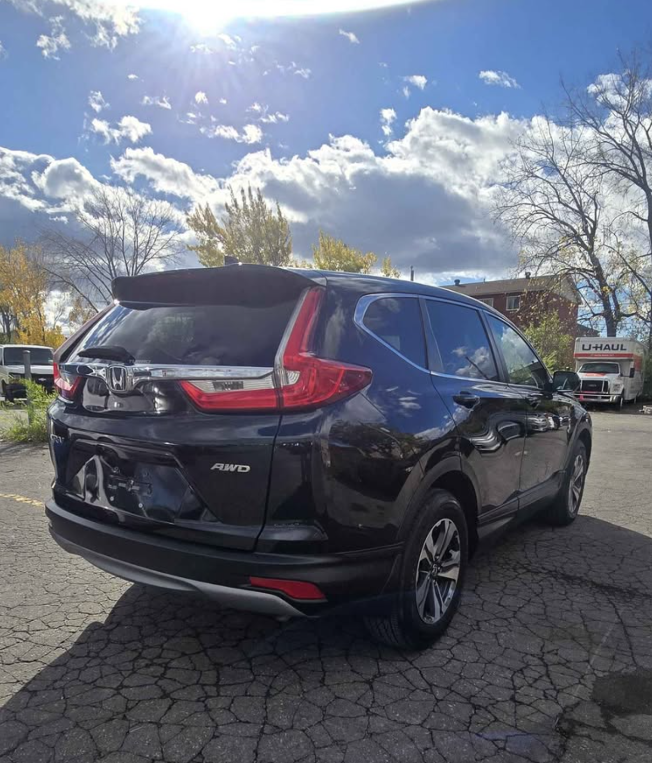 Black Honda CR-V SUV parked on a cracked asphalt lot with trees and clouds in the sky, and a U-Haul truck in the background.
