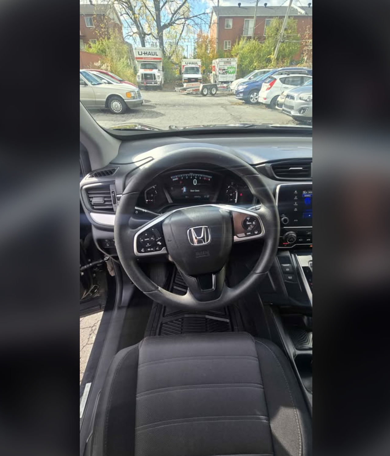 View from inside a Honda vehicle showing the steering wheel, dashboard, and front seat, with a parking lot and U-Haul trucks visible through the windshield.