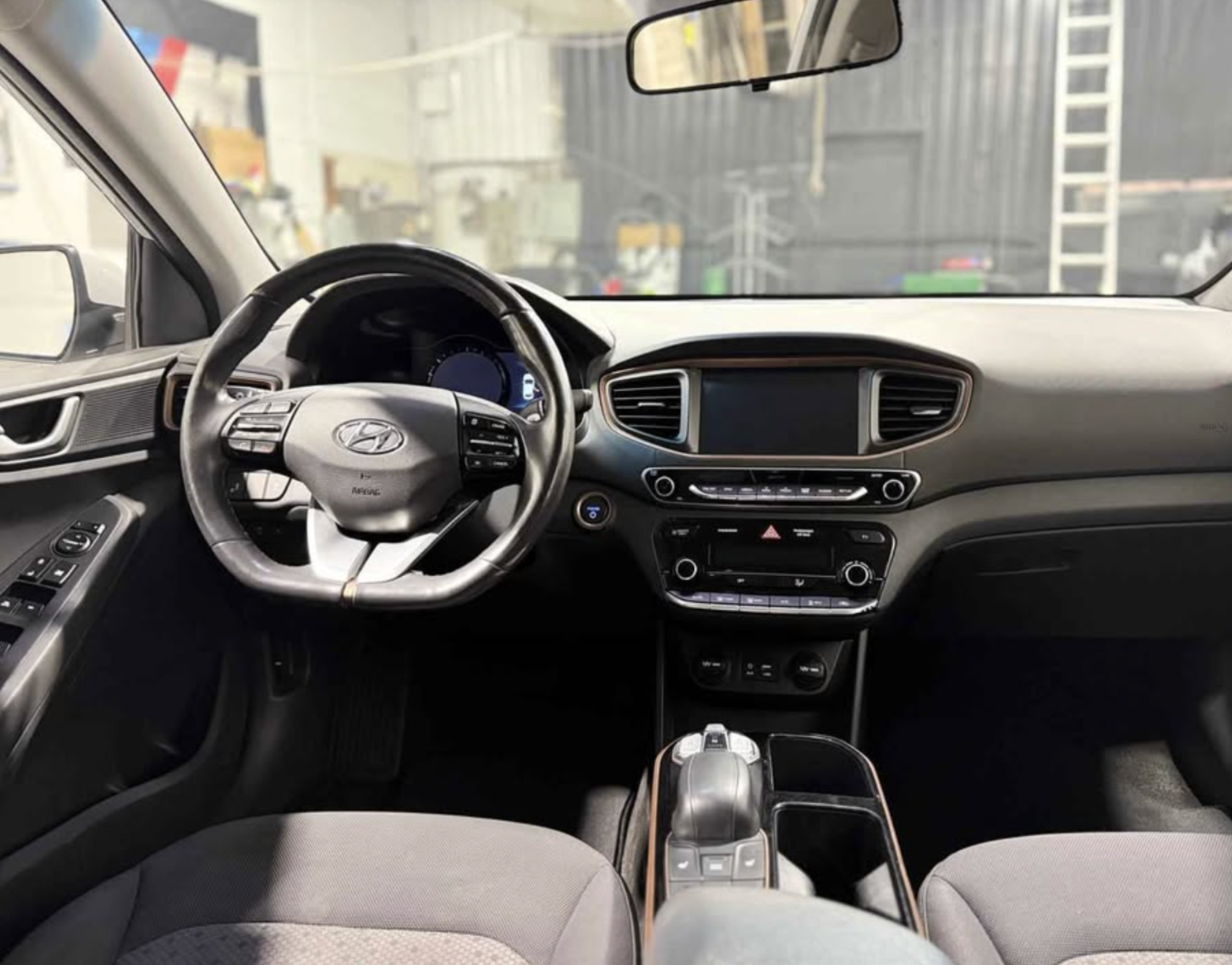 Photo of a car interior showing the steering wheel, dashboard, and center console. The steering wheel has a Hyundai logo, and the dashboard includes a touchscreen display and climate controls. The view is from the front seats looking towards the fron