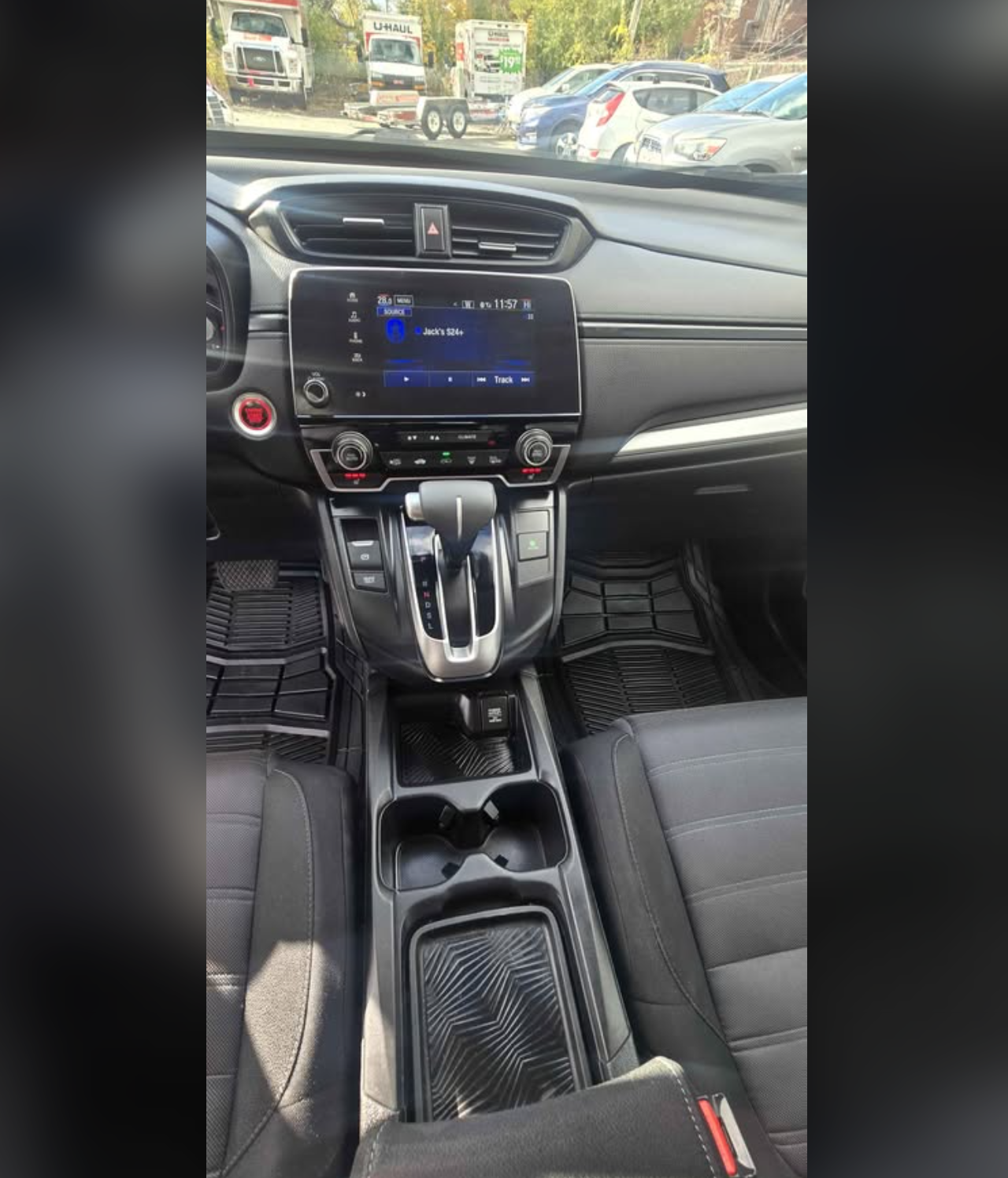 The interior of a modern vehicle showing the dashboard, infotainment screen displaying a song, gear shift, and center console with cup holders, with parked cars and trucks visible through the windshield.