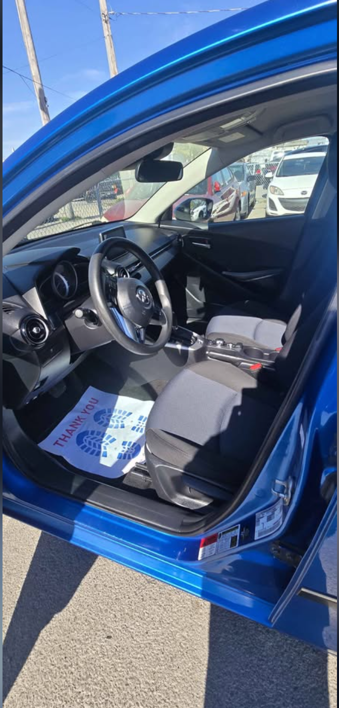 Interior of a blue car showing the steering wheel, dashboard, and front seats, with a paper on the driver's side floor mat that reads 'Thank You' with a blue footprint design.