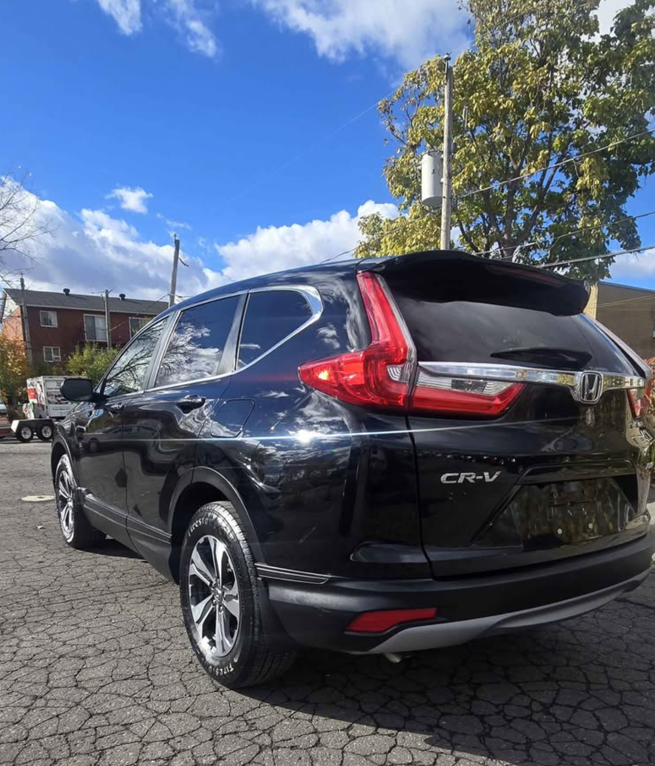 Black Honda CR-V parked on a cracked asphalt surface with trees, houses, utility poles, and a partly cloudy blue sky in the background.