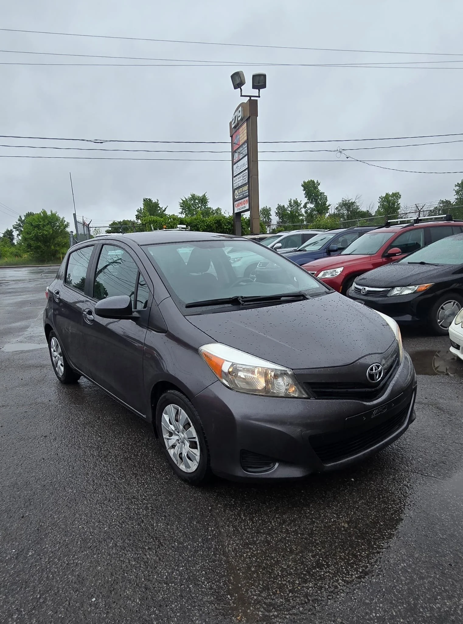 Gray Toyota hatchback parked in a wet car lot on a rainy day, with other vehicles and a signboard in the background.