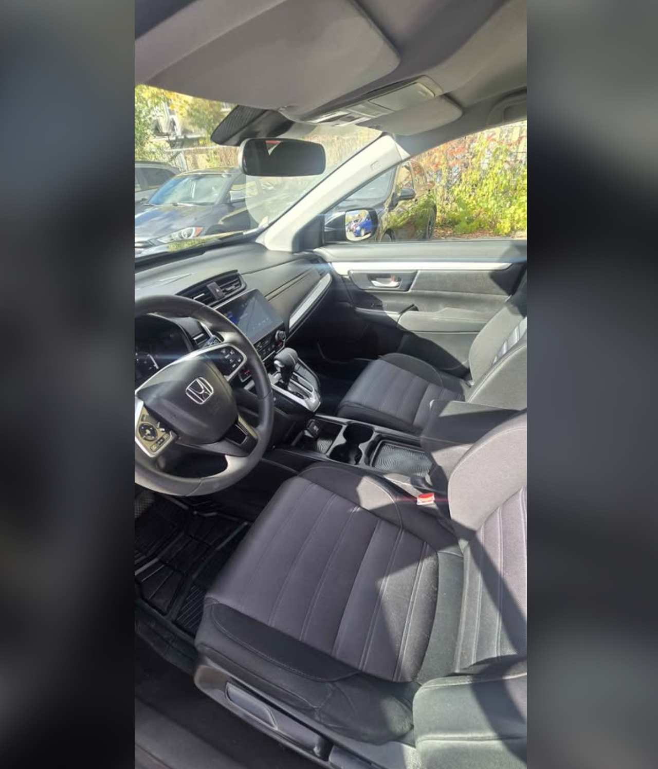 Interior view of a Honda vehicle showing the driver's seat, steering wheel, dashboard, center console, and front passenger seat, with a view through the windshield of parked cars and trees outside.