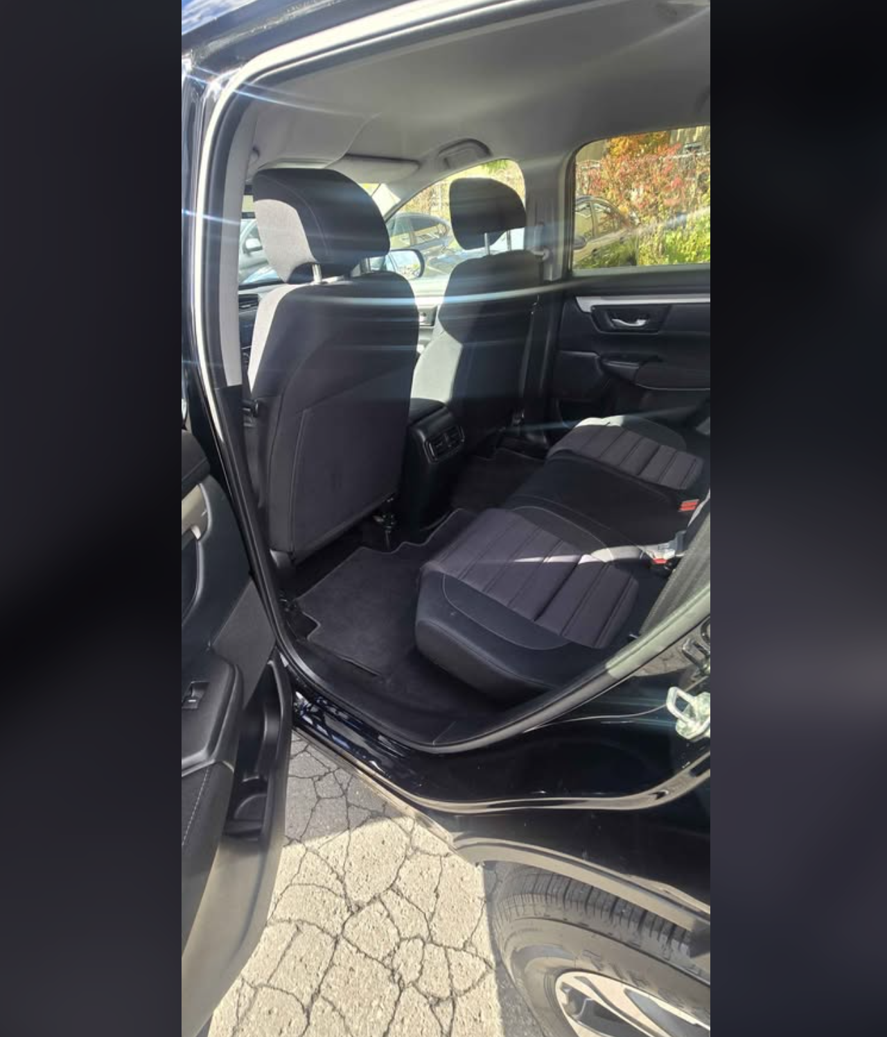 The interior of a black SUV, showing the backseat area with black fabric seats, front seats, and the rear door open. Outside, there are cars and trees with fall foliage visible through the windows.