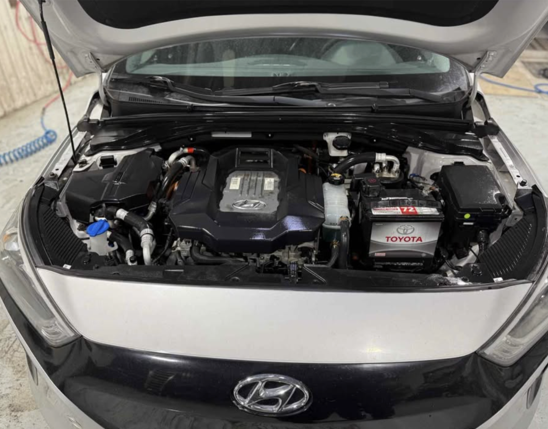 Under the open hood of a silver Hyundai vehicle, the engine bay reveals various components including the Hyundai engine cover, Toyota battery, and other engine parts.