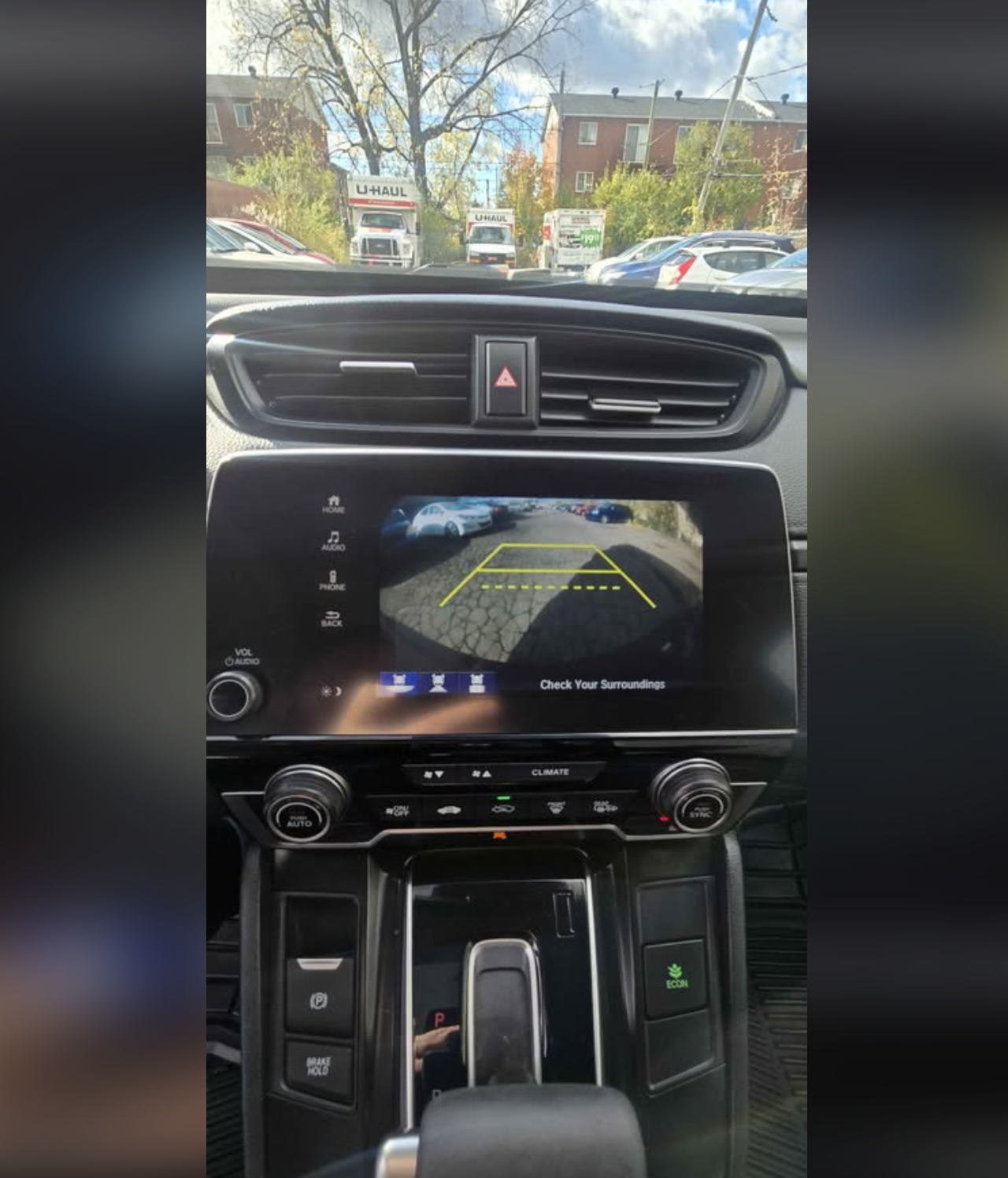 View from inside a car showing the dashboard with a rearview camera display. The display shows the parking lot behind, with colored guide lines and a warning message to check surroundings. Outside, U-Haul trucks are parked or moving, trees and buildi