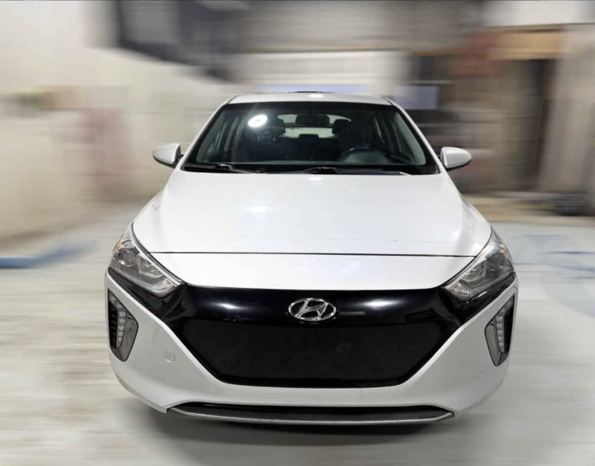 Front view of a white Hyundai car with a black grille, parked in a garage with a blurred background.