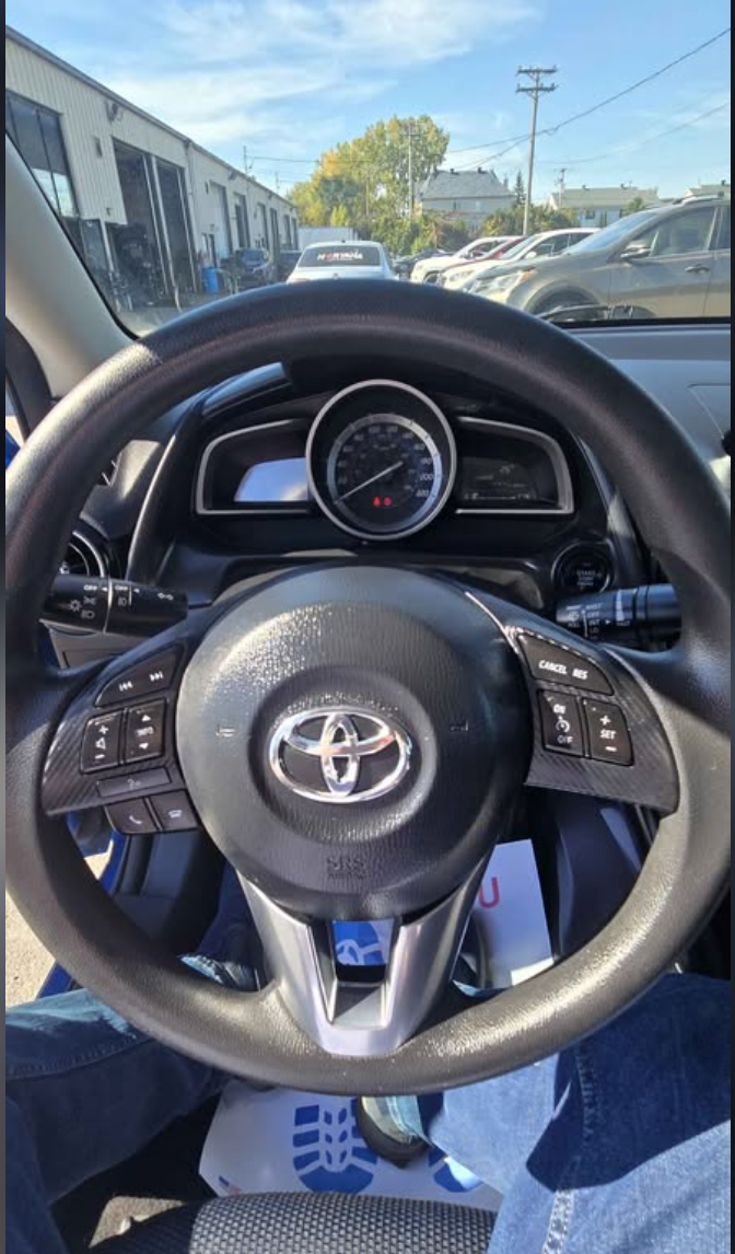 Inside view of a Toyota car dashboard and steering wheel, with a parking lot in the background, including parked cars and a building.