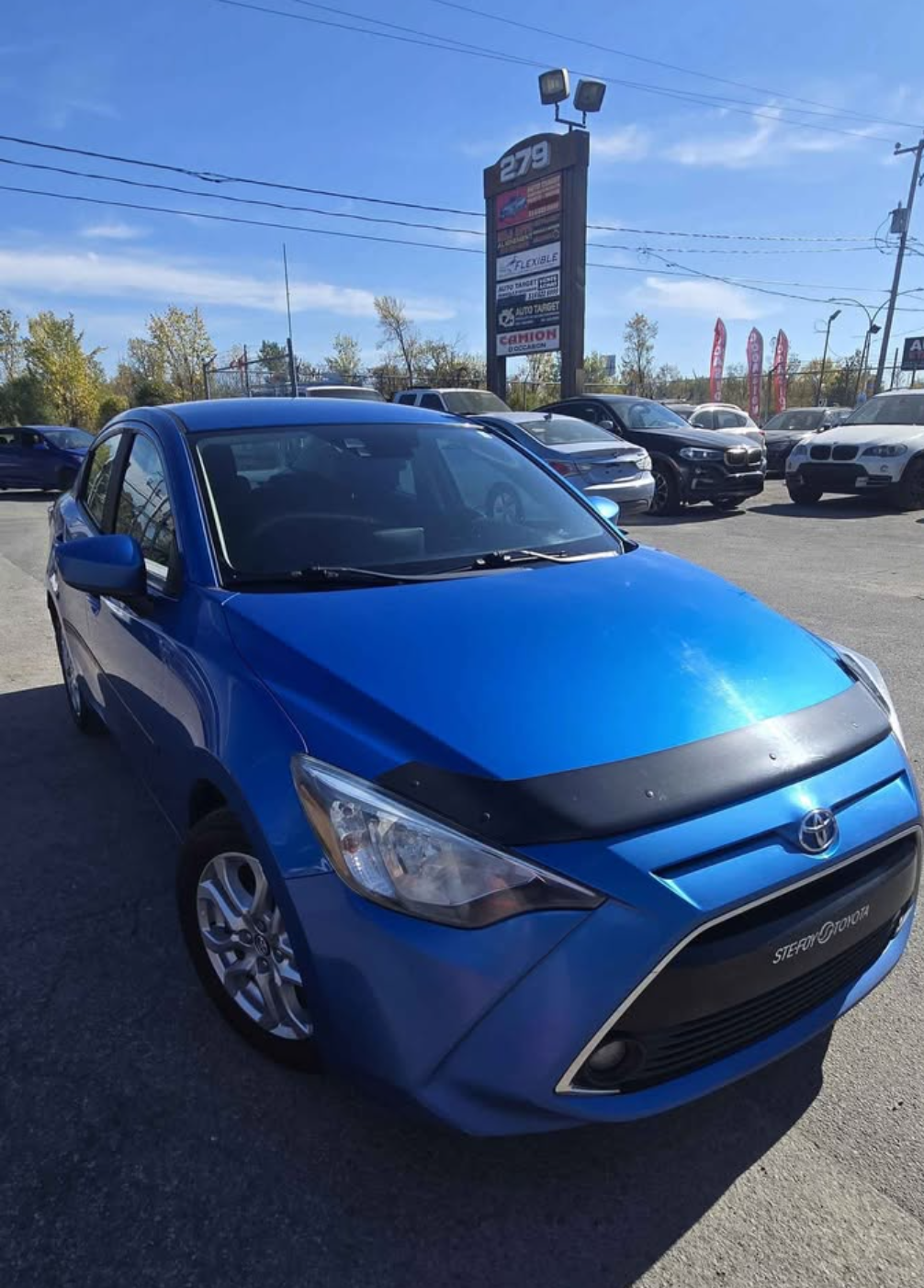 Blue Toyota sedan parked in a dealership lot with other cars in background and a tall sign showing various auto parts stores.