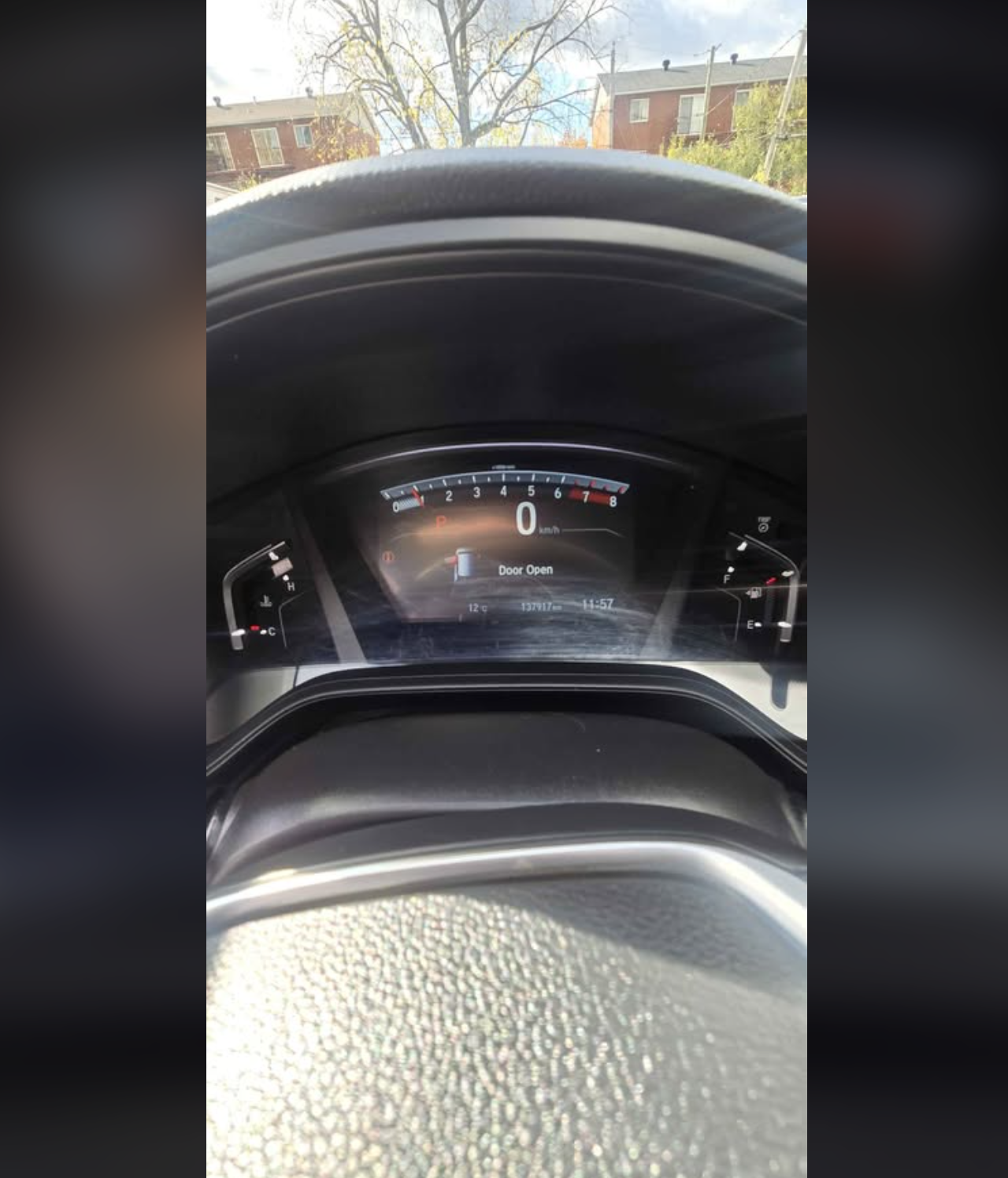 Car dashboard showing 0 km/h speed, open door warning, and outside view of residential buildings and trees.