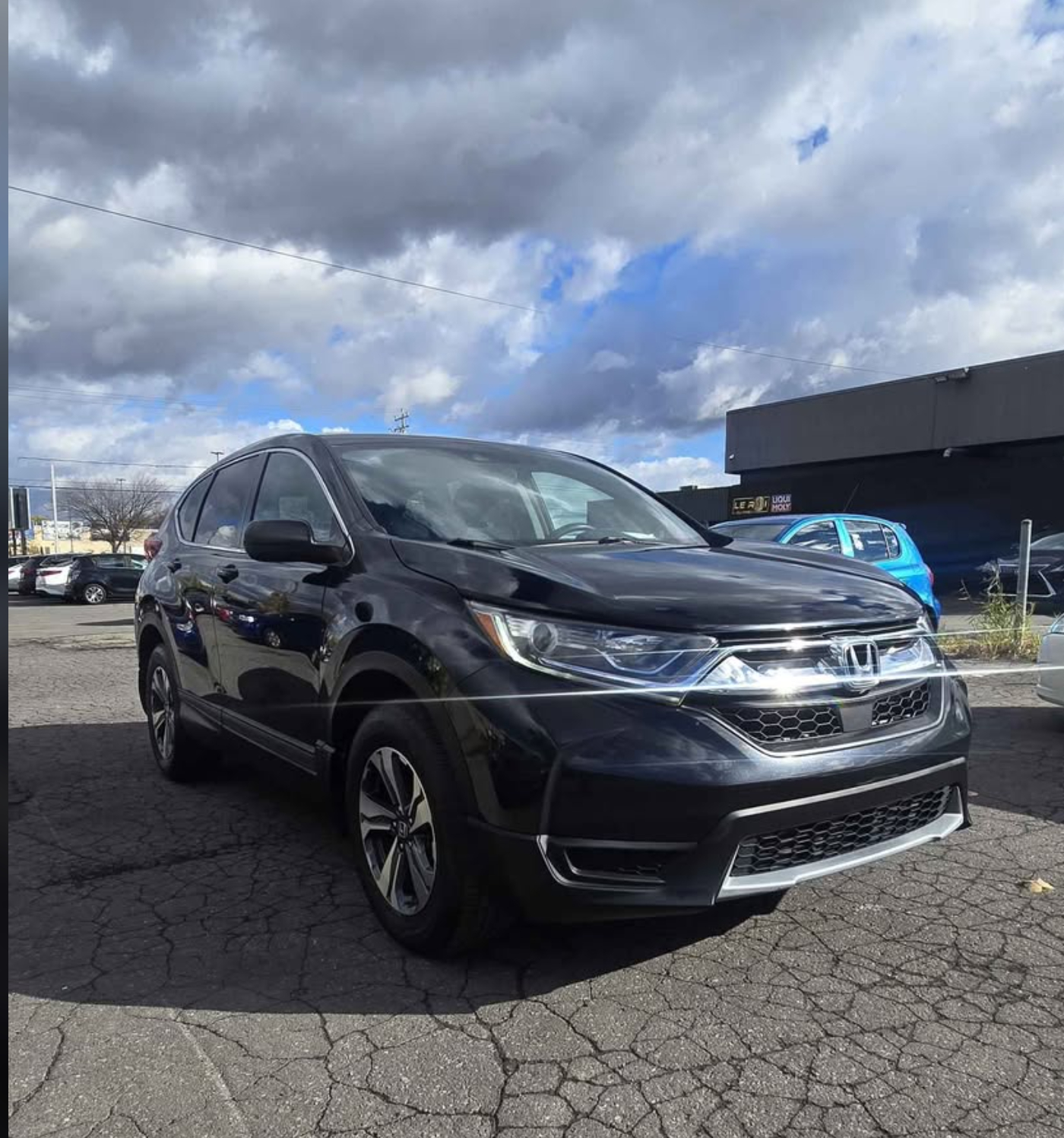 Black Honda CR-V parked on cracked asphalt in a parking lot with other vehicles and a store building in the background under cloudy sky.