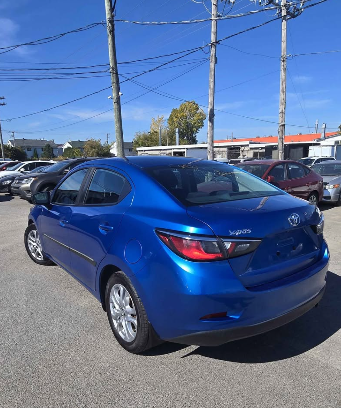 Blue Toyota Yaris parked in a parking lot with other cars, utility poles, and power lines overhead, under a clear blue sky.