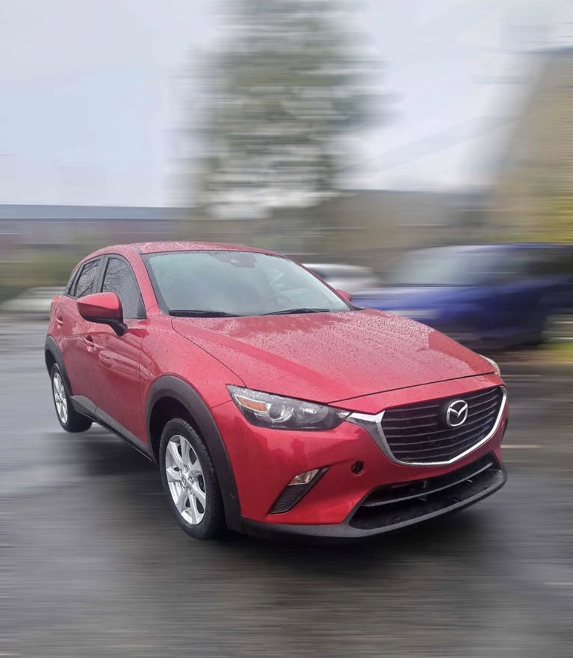 Red Mazda CX-3 driving on a wet road, with a blurred background, possibly on a rainy day.