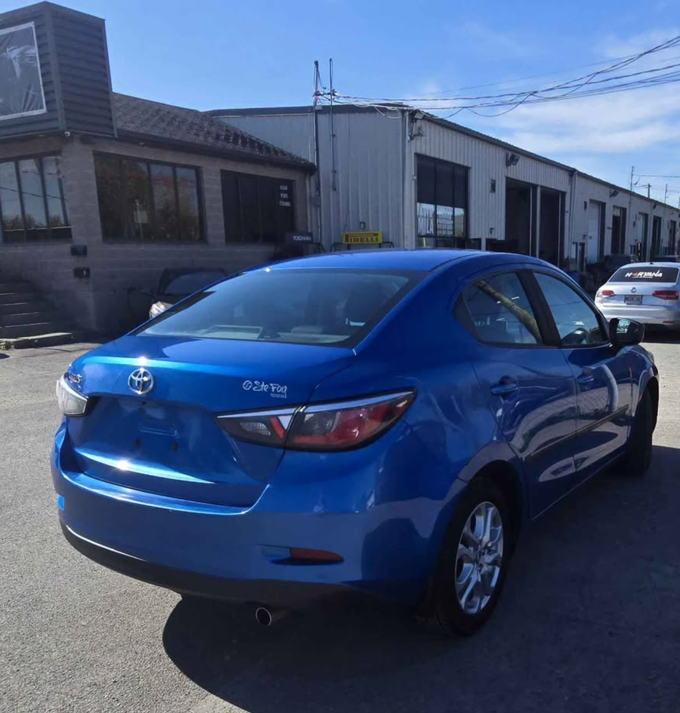 Blue Toyota car parked outside a building with industrial-style exterior, with other cars and power lines in the background on a sunny day.