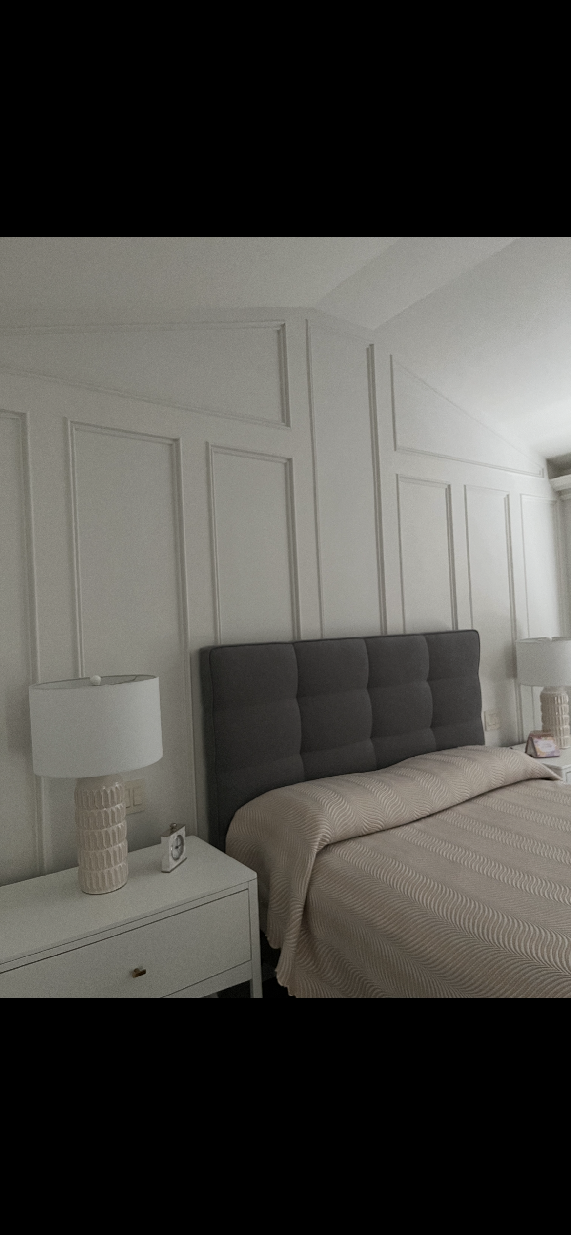 A bedroom with white paneled walls, a dark gray upholstered headboard, a cream-colored bedspread with a subtle pattern, and two white nightstands with beige textured lamps and small picture frames.