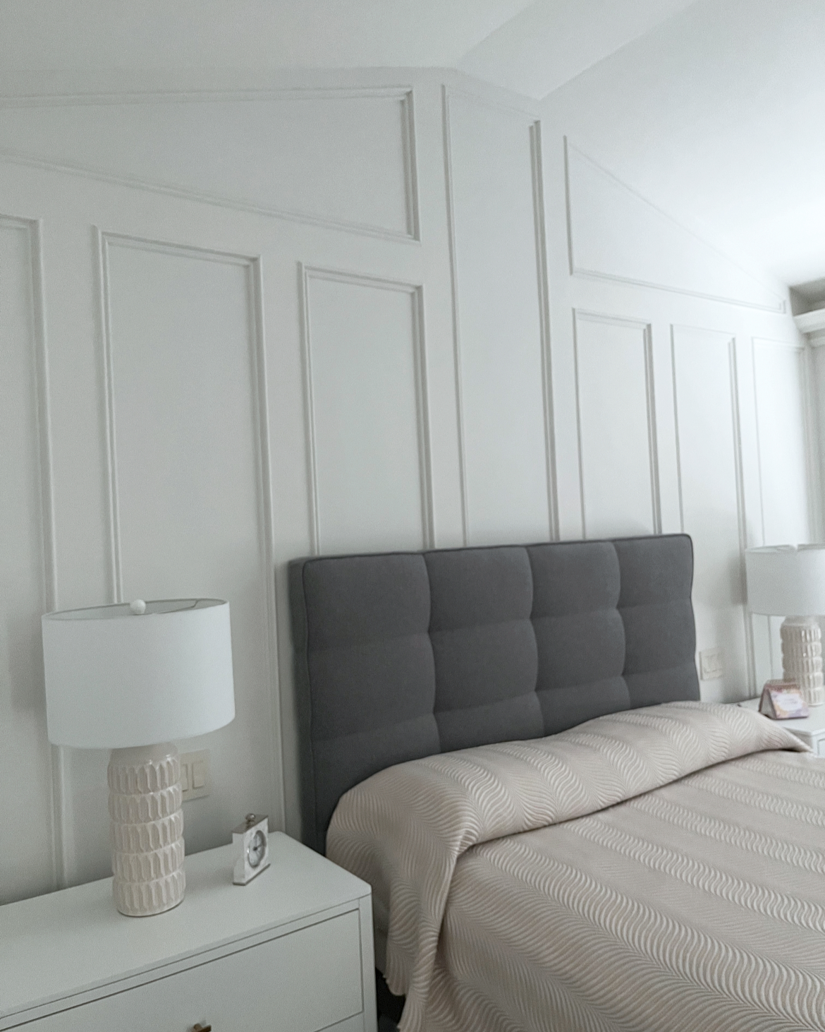 A bedroom with a gray upholstered headboard, white side tables with lamps, and a bed with beige and white patterned bedding, white paneled walls, and a window with curtains.