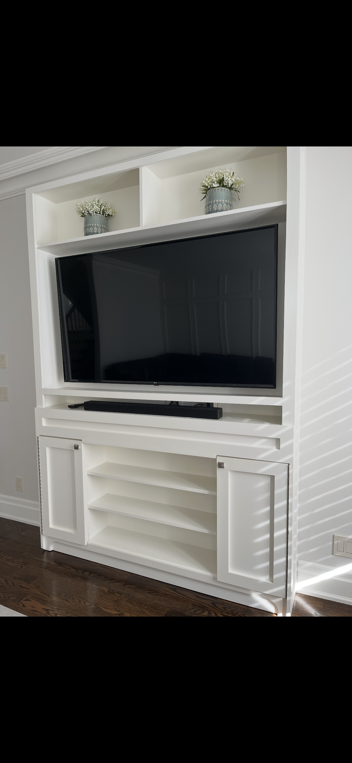 White entertainment center with open shelves and a black flat-screen TV mounted on it, with two decorative flower vases on the top shelf and a soundbar below the TV, located on a hardwood floor.