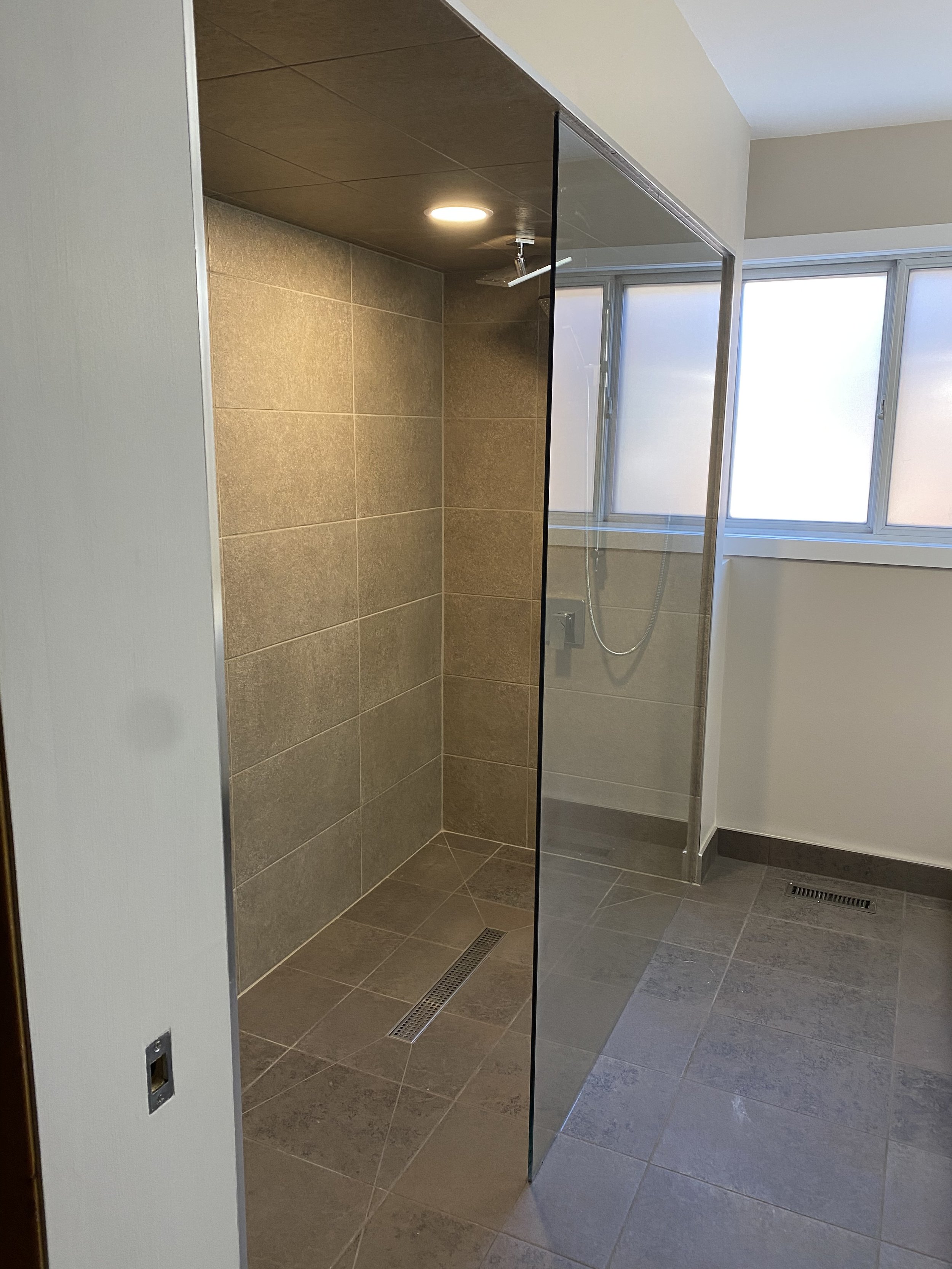 Empty modern shower with tiled walls and floor, glass door, overhead light, and frosted windows.