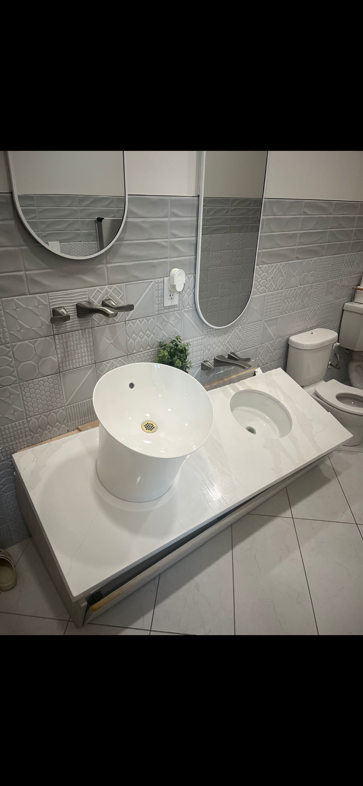 Modern bathroom with patterned gray tiles, a white marble countertop with a round vessel sink and an alcove for a smaller sink, two mirrors, a small plant, and a toilet.