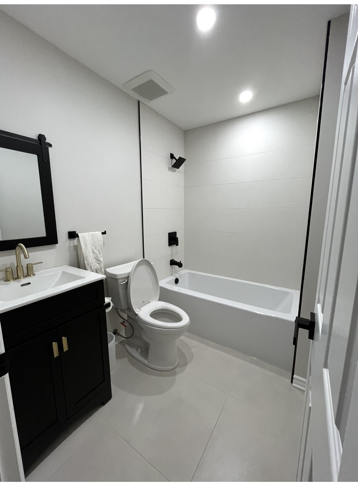 Modern bathroom with a black vanity, white sink, gold faucet, toilet, and bathtub with black fixtures. White walls, gray tile floor, ceiling lights, and black accents.