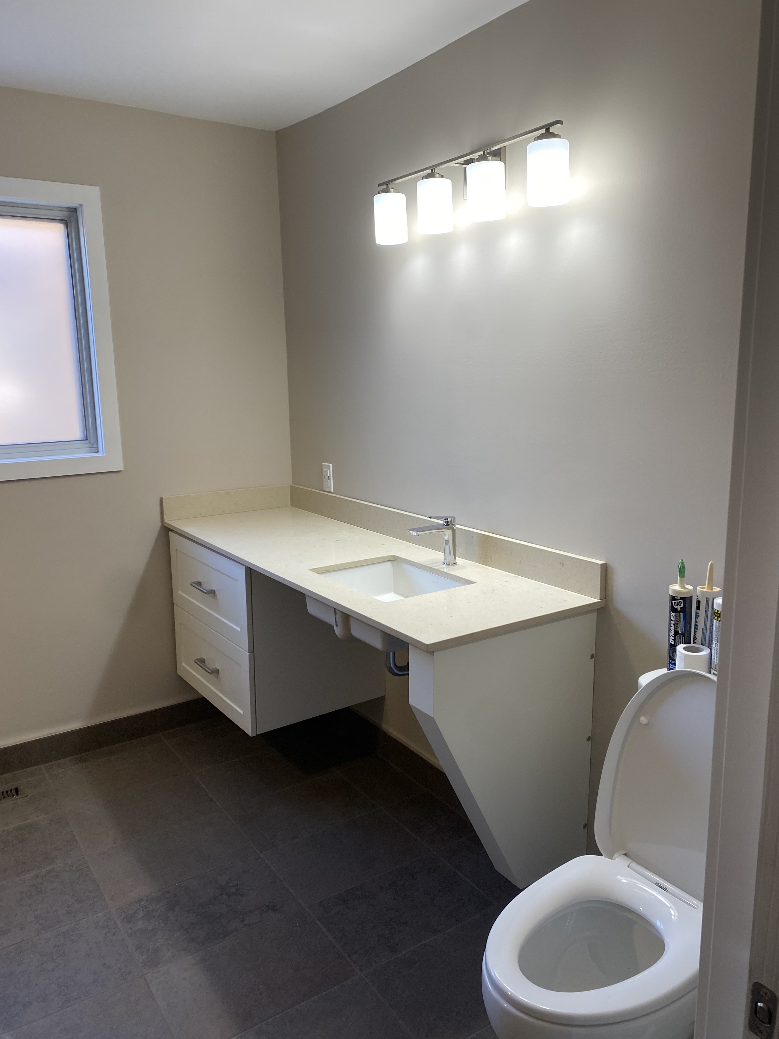 A laundry room with beige walls, a white countertop with a small integrated sink, and a cabinet with drawers beneath. There is a window on the left, overhead lighting fixture with four bulbs, and a toilet on the right side with toilet paper and caulking tubes nearby.