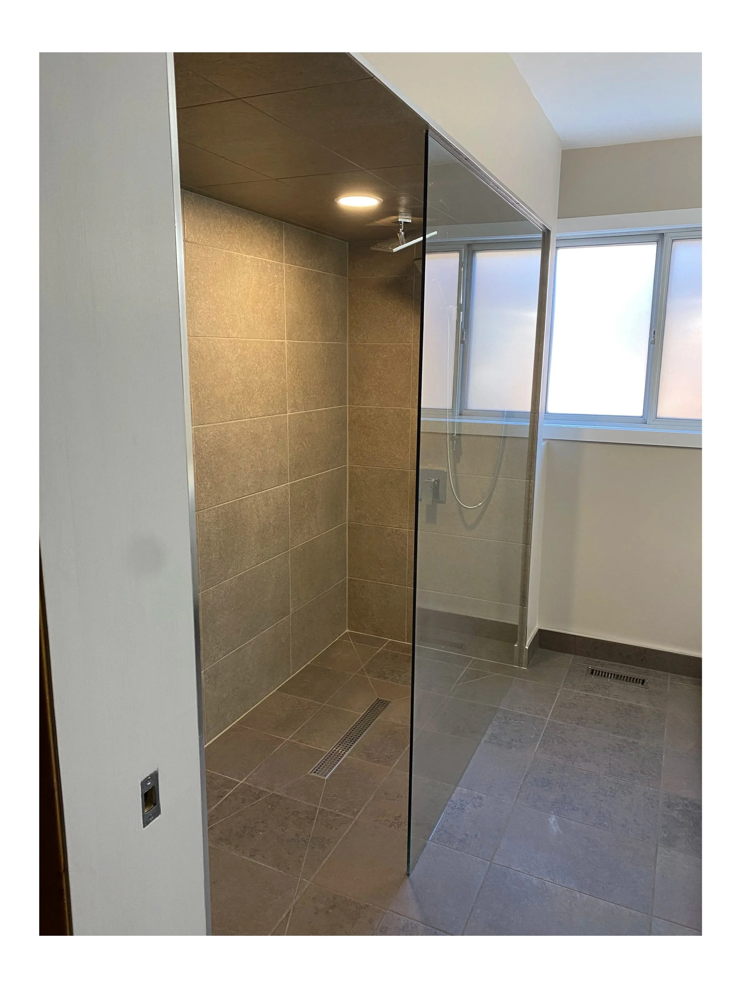 Empty modern shower with a glass partition, beige tiled walls, gray tiled floor, ceiling light, and frosted windows