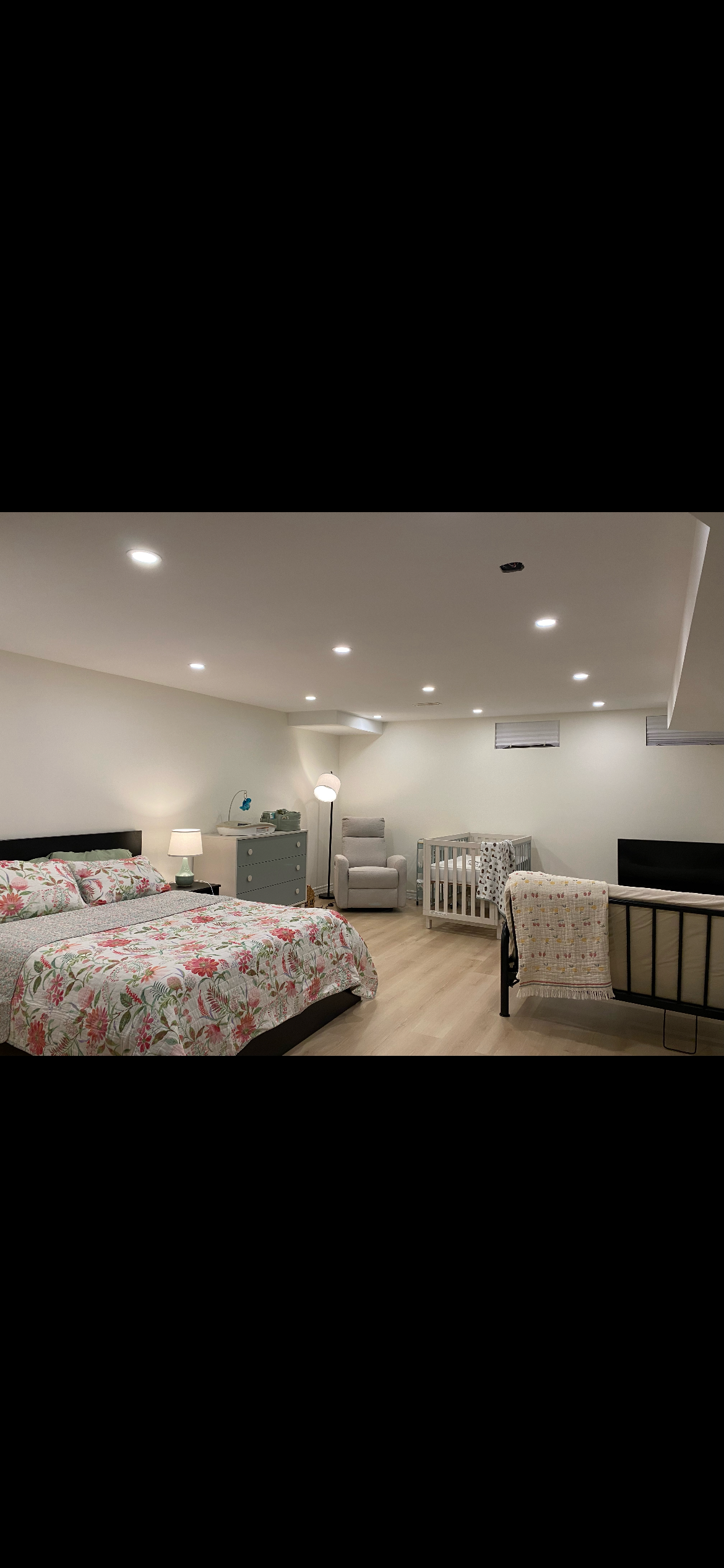 A bedroom with a bed covered in floral bedding, a nightstand with a lamp, a light blue dresser with items on top, a floor lamp, a white armchair, a crib, a changing table, and a black metal bed frame in a well-lit room with white walls and hardwood flooring.