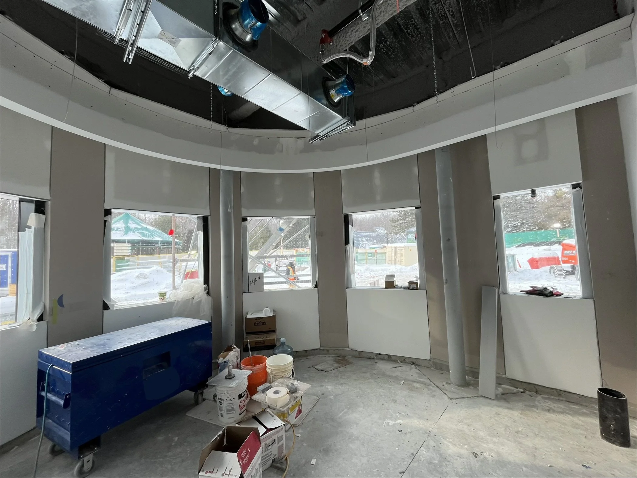 Interior of a building under construction with multiple windows, unfinished walls, construction materials, and equipment scattered on the floor.