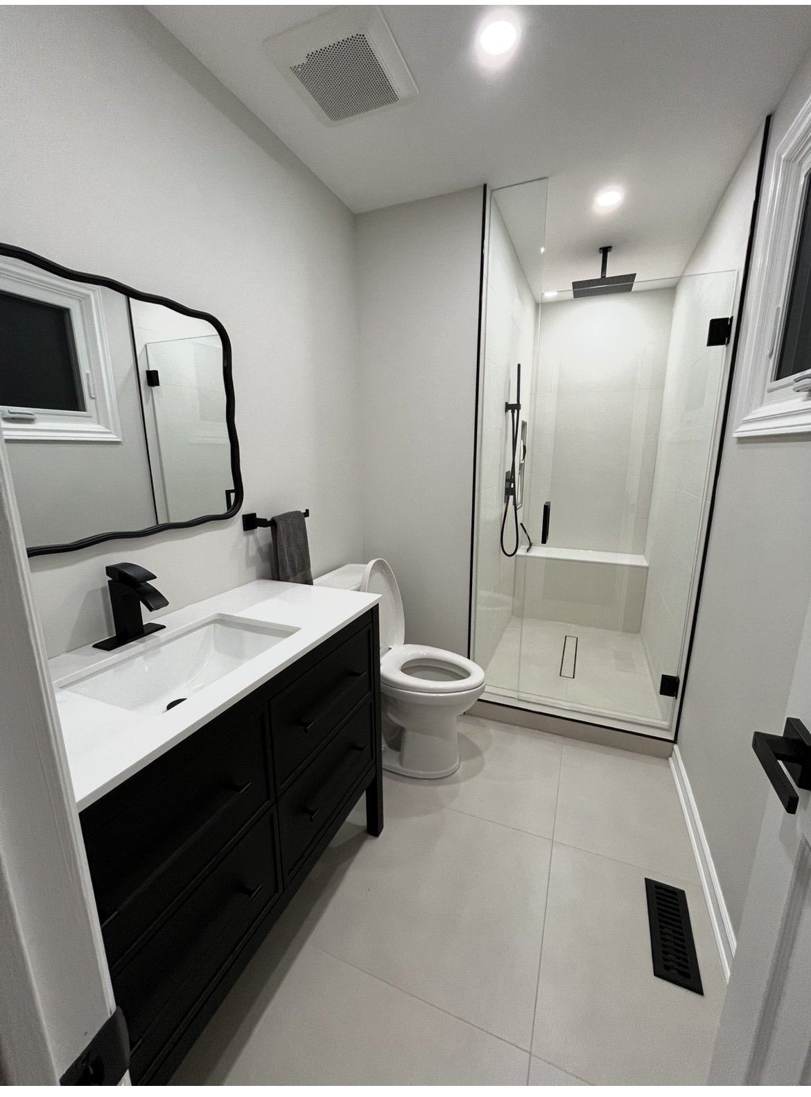 Modern bathroom with white walls, a black vanity with a white countertop, and a black faucet. There is a mirror above the sink, a towel hanging on a black towel holder, and a toilet next to the vanity. A glass-walled shower with a rainfall showerhead and a handheld showerhead is at the back of the room. The bathroom has a window, a vent on the ceiling, and a floor vent.
