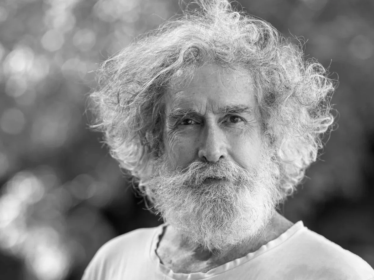 Black and white photo of man with soft, grey hair, looking into camera, wearing white t-shirt